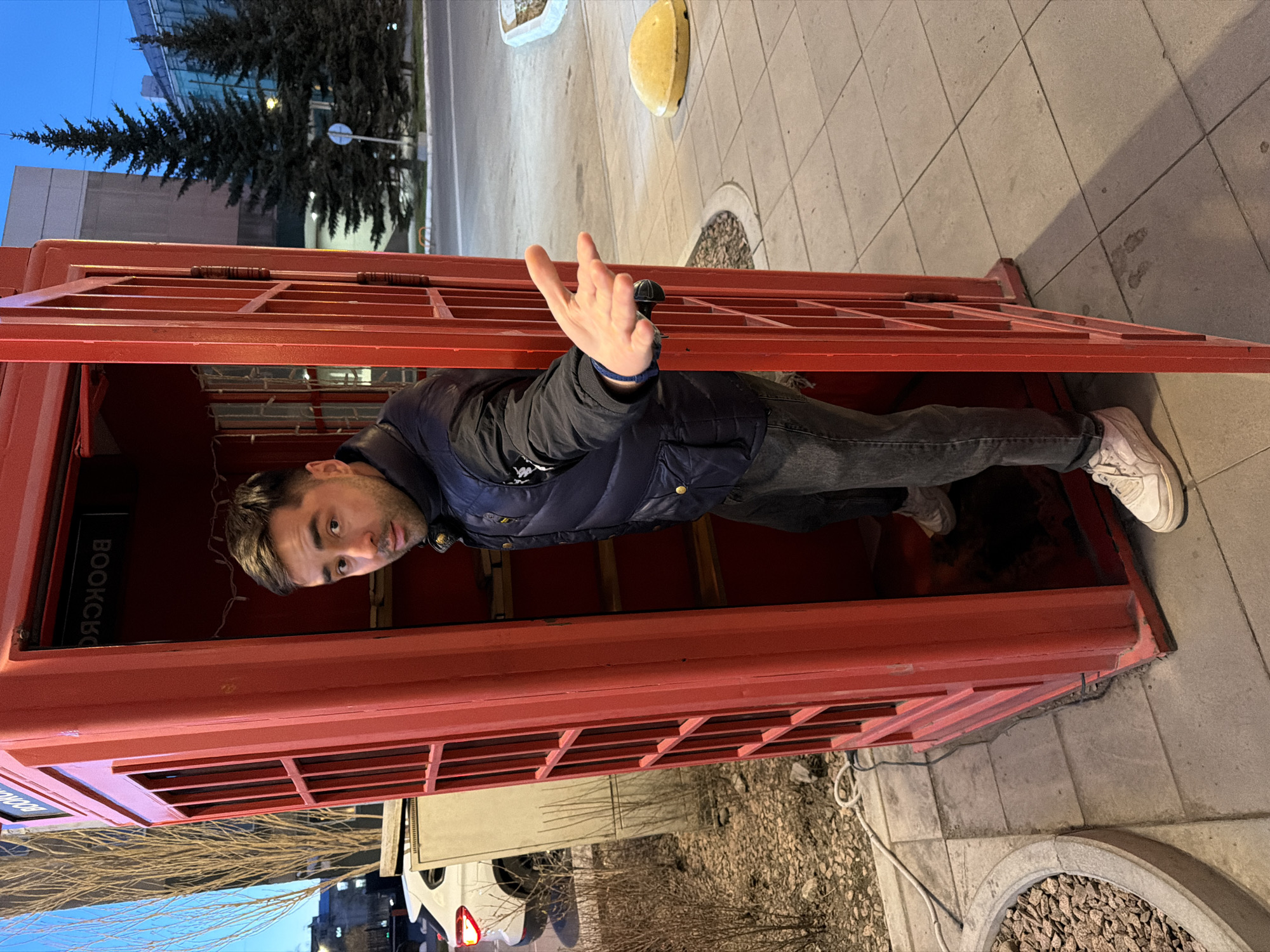 A man is inside a red telephone booth. He is wearing a dark blue puffer jacket, grey jeans, and white sneakers. He is looking toward the camera and holding up his hand. The telephone booth has a sign that reads "BookCrossing" vertically. Outside the booth is a sidewalk with grey paving stones. In the background, there is a street, trees, and a building. This is a location in the city of Krasnoyarsk, Russia.
