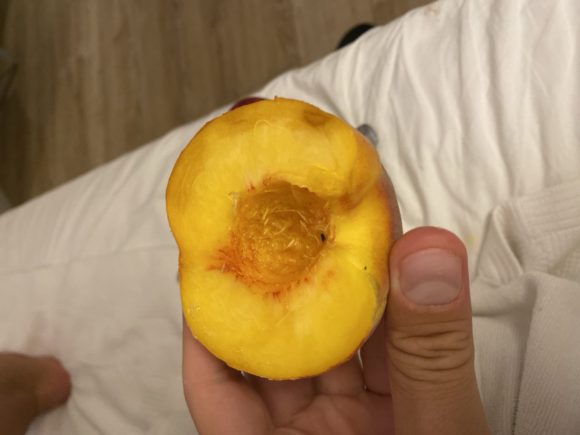 A close-up, eye-level shot shows a hand holding a halved peach. The peach is bright yellow with red tinges near the pit cavity. The hand, with visible fingerprints and a clean fingernail, is gripping the peach from the bottom left. The background consists of white fabric, likely bedding, and a wooden floor. A person's foot is partially visible in the upper left corner. The scene appears to be indoors, possibly in a bedroom or hotel room. There is no indication of the city of Aksu, Turkey, in the image.
