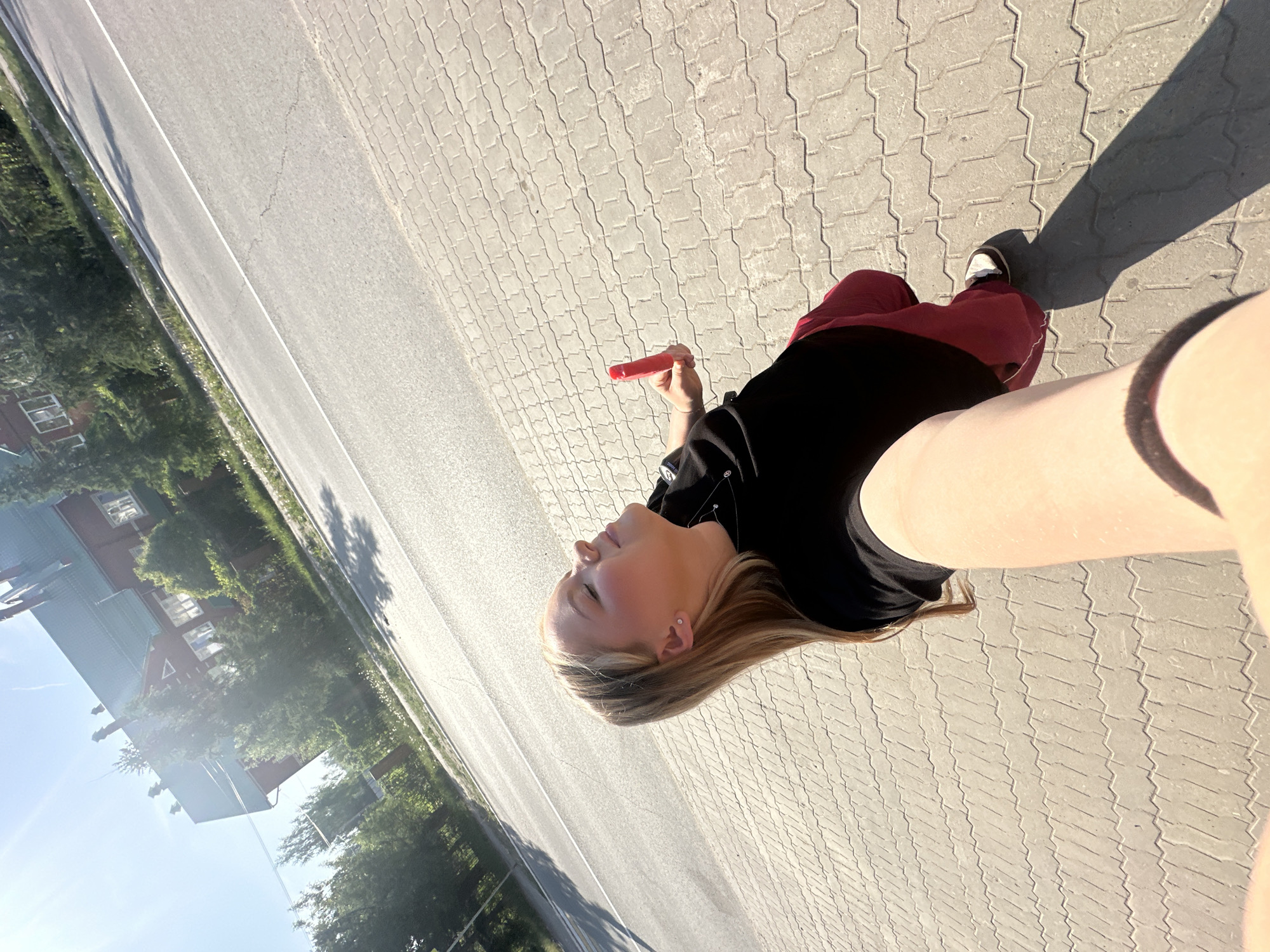 A woman is holding a red popsicle in the city of Chaikovsky, Russia. She is on a sidewalk with patterned concrete pavers. She is wearing a black shirt and maroon pants, and one shoe is visible. A street and buildings are in the background. The lighting suggests a sunny day. The picture appears to be a selfie taken at a low angle.