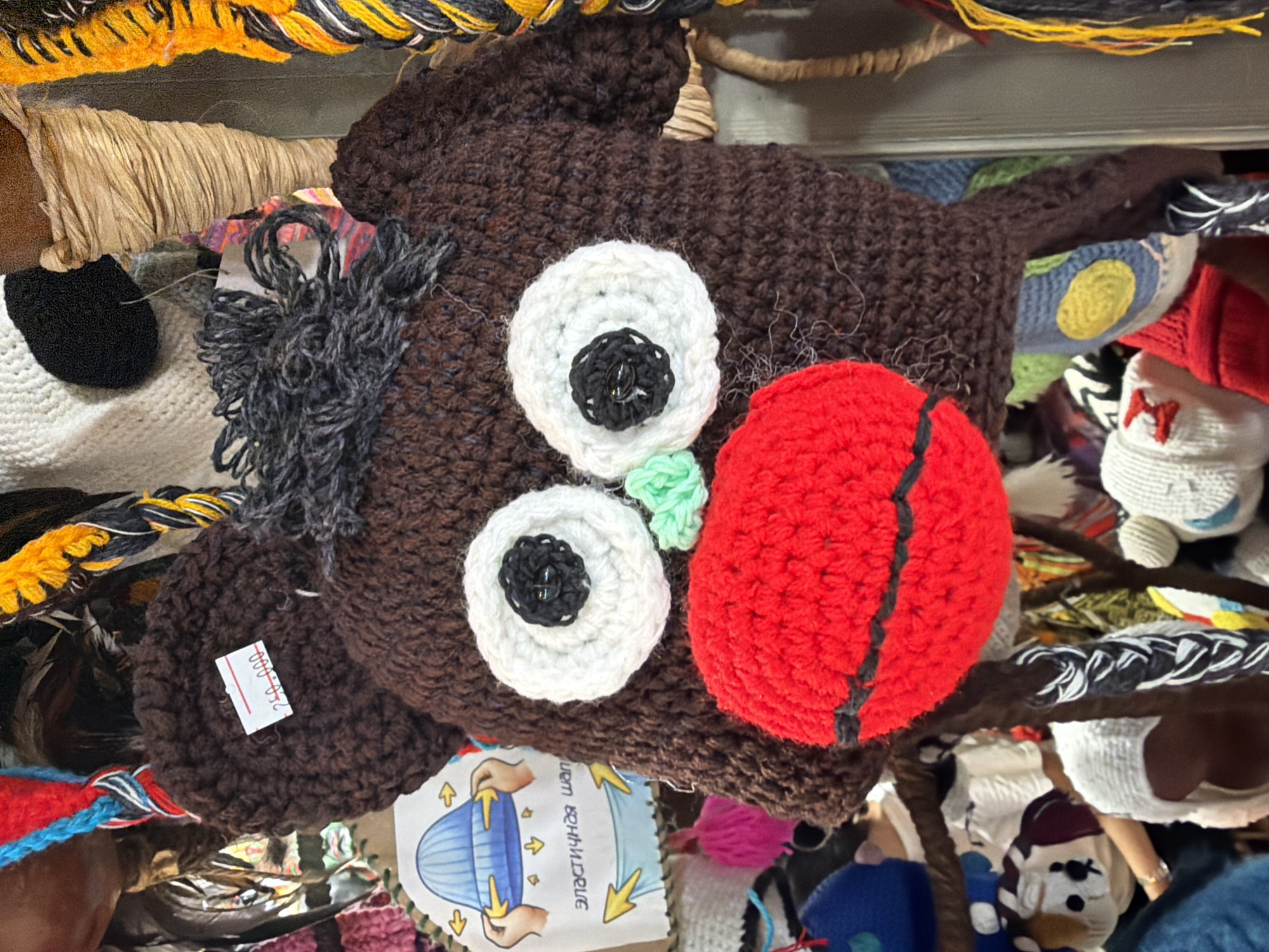 The image shows a close-up of a brown knitted hat with exaggerated facial features, resembling a monkey or a bear. It has large, white crocheted eyes with black button pupils, a bright red crocheted mouth with a black stripe, and small green crocheted nostrils. Dark gray yarn forms the hair on top of the head. A white price tag with red writing, reading "250.000", is attached to the hat. The hat is part of a display of various knitted items, including other hats with different designs and colors, and woven materials in the background. The setting appears to be a market stall in Nha Trang Ward, Vietnam, given the presence of the price tag in Vietnamese currency and the overall aesthetic of handicraft sales. The lighting is diffused, suggesting an indoor or shaded outdoor environment.