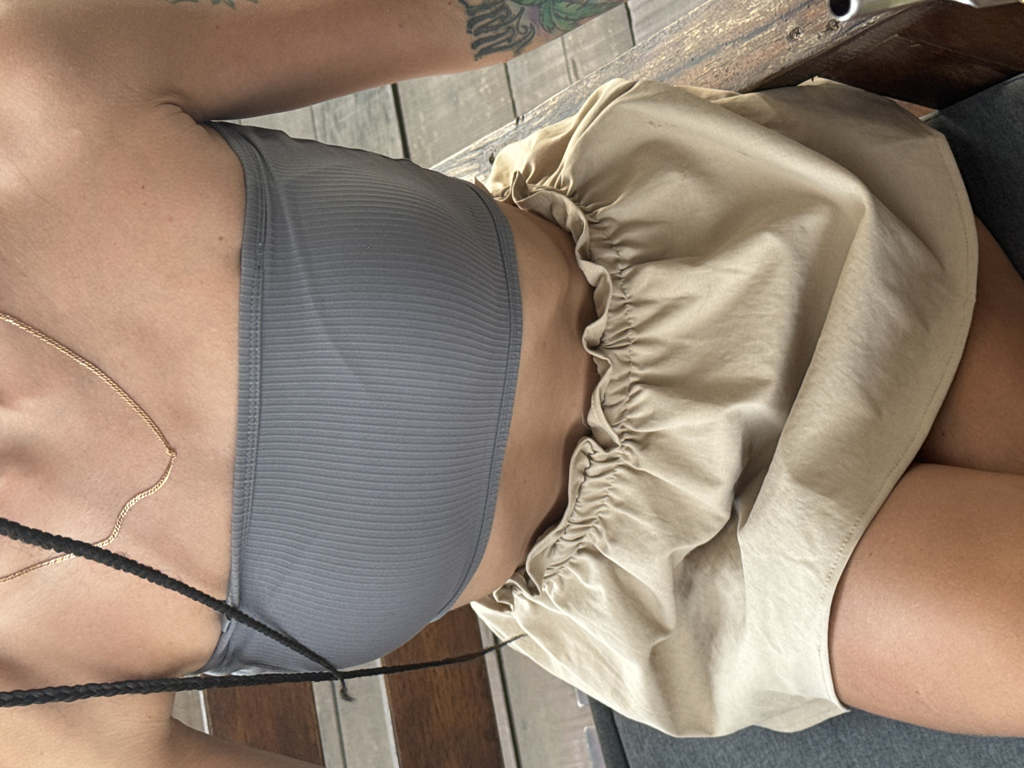 The image shows a close-up, top-down view of a person seated, likely in an outdoor or semi-outdoor setting within Hà Giang 1 Ward, Vietnam. The subject, visible from the upper chest to the upper thighs, wears a grey ribbed crop top and a light beige or khaki-colored skirt with a ruffled, elasticated waist. A thin gold chain necklace is visible around their neck, and strands of dark braided hair extend across their chest. A tattoo featuring green and black ink, possibly depicting foliage or an abstract design, is visible on their upper right arm.
The person is seated on a grey surface, possibly a cushion or upholstered chair. Behind and to the side of the subject, a wooden structure, likely a table or bench, with visible grain and fasteners, suggests a rustic or natural environment. The bright, even lighting indicates it is daytime. The activity captured is simply sitting. No visible text is present in the image.