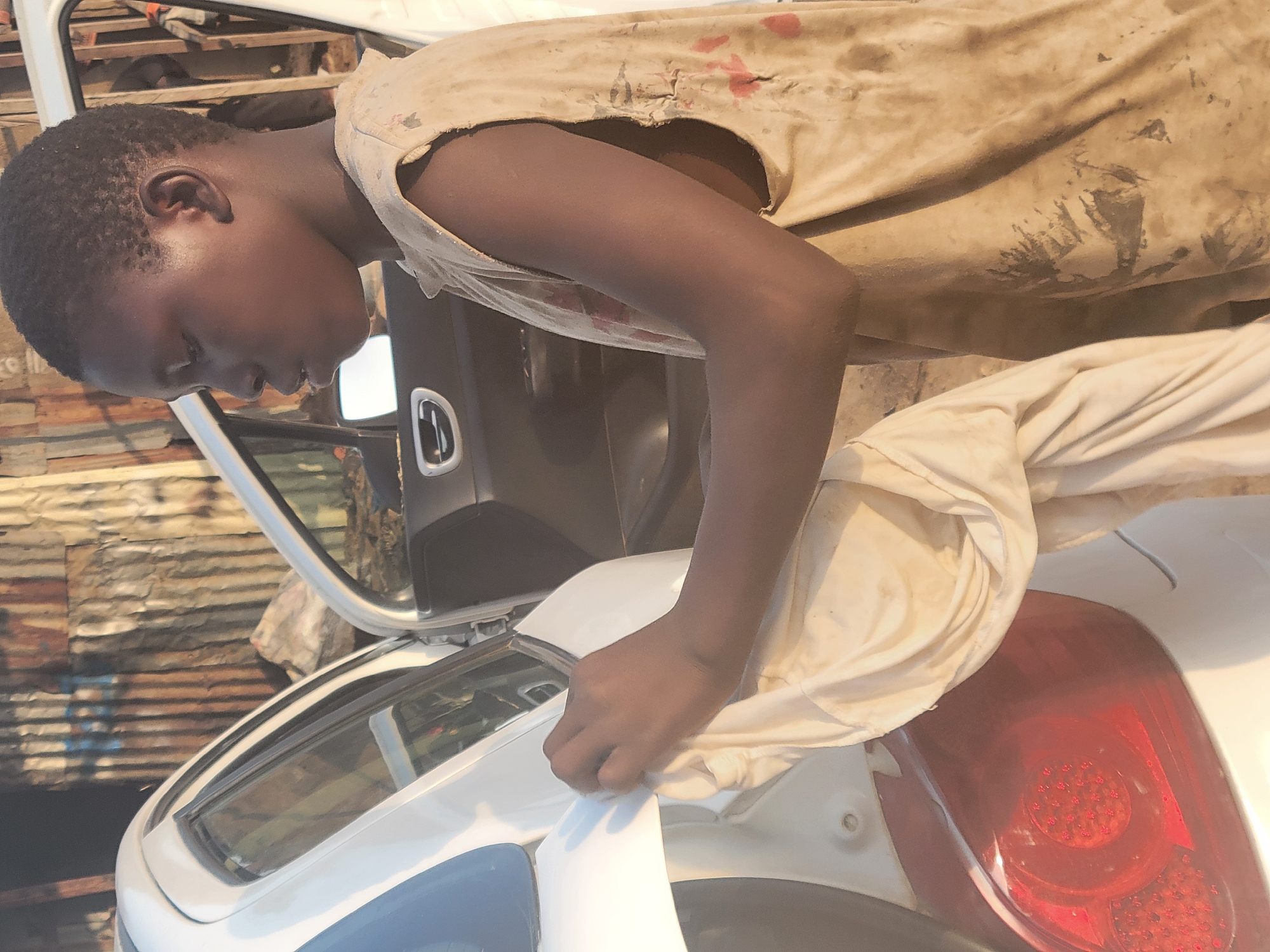 A young, dark-skinned person in Kwamba, Nigeria, is polishing the rear of a white car with a light-colored cloth. The person wears a tattered, stained beige tank top and light-colored pants. The car's open door suggests it might be undergoing cleaning or repair. The background includes corrugated metal sheets, indicating a simple structure, and sunlight is illuminating the scene.