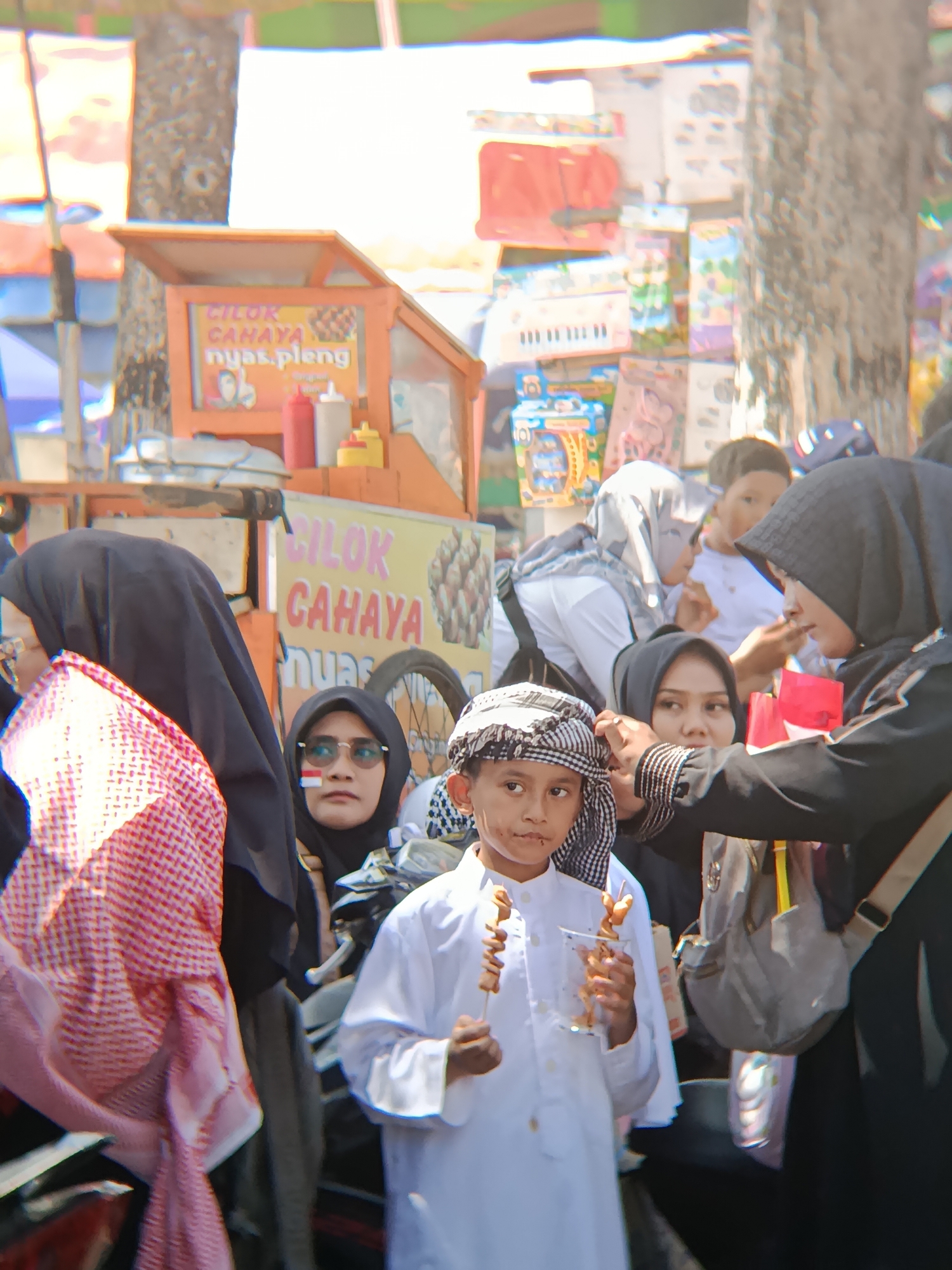 In Desa Kebon Manis, Kecamatan Cilacap Utara, Indonesia, a group of people are gathered outdoors, possibly at a street market or festival. In the foreground, a young boy wearing a white tunic and a checkered keffiyeh is holding two skewers of small, browned food items, appearing to be sausages or meatballs, in a plastic cup. He is looking directly at the camera. To his right, a woman wearing a black hijab is adjusting his keffiyeh. Behind them, other individuals are visible, including a woman in a black hijab with sunglasses and a red and white Indonesian flag painted on her cheek, and another person wearing a light-colored hijab. In the background, a food cart with signage that reads "CILOK CAHAYA" is present, along with various colorful items, potentially merchandise or decorations, displayed on shelves. The environment appears bright and sunny, with trees visible.