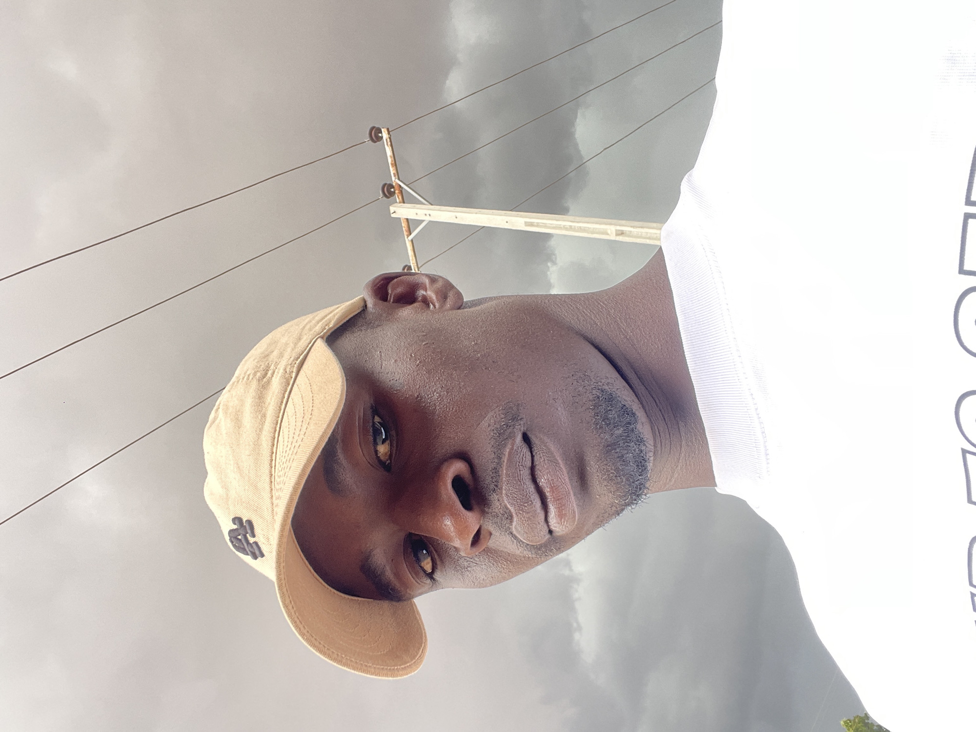 A medium, portrait-style outdoor shot from a low angle captures a dark-skinned man wearing a tan baseball cap, posed against a cloudy sky. The man is facing the camera with a neutral expression. Utility poles with electrical lines extend into the sky behind him, typical of urban environments. Given the prompt, this image is likely taken in the city of Jos, Nigeria. The man is wearing a white T-shirt, the partial view of which suggests there may be text on the shirt.