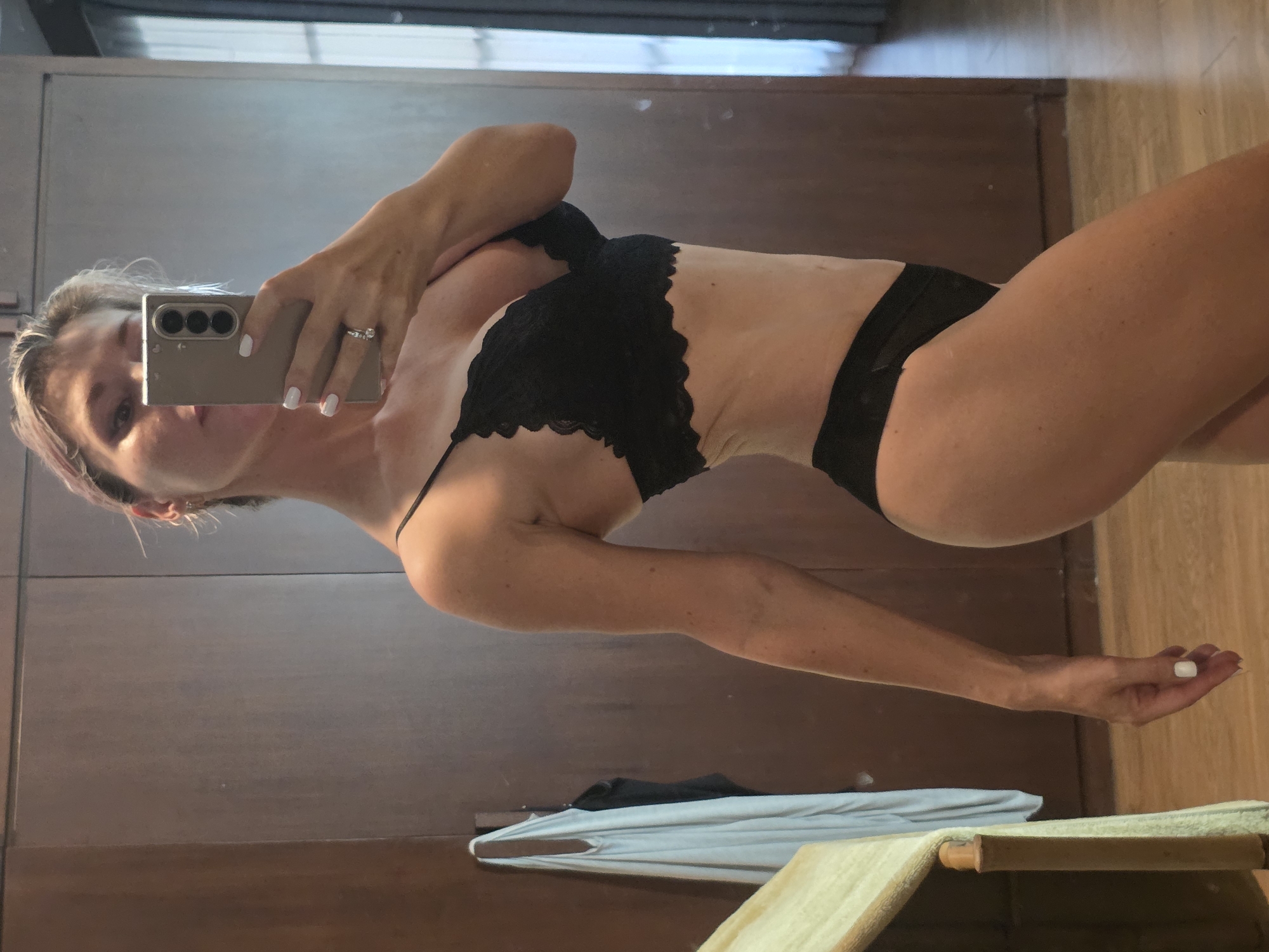 A woman is taking a mirror selfie in a room with wooden floors and a large wooden closet. She is wearing black lingerie, a black bra with lace detailing, and black underwear. Her right hand is holding a silver smartphone, and she is wearing a ring on her left hand. A light blue garment hangs on a hook on the closet door.