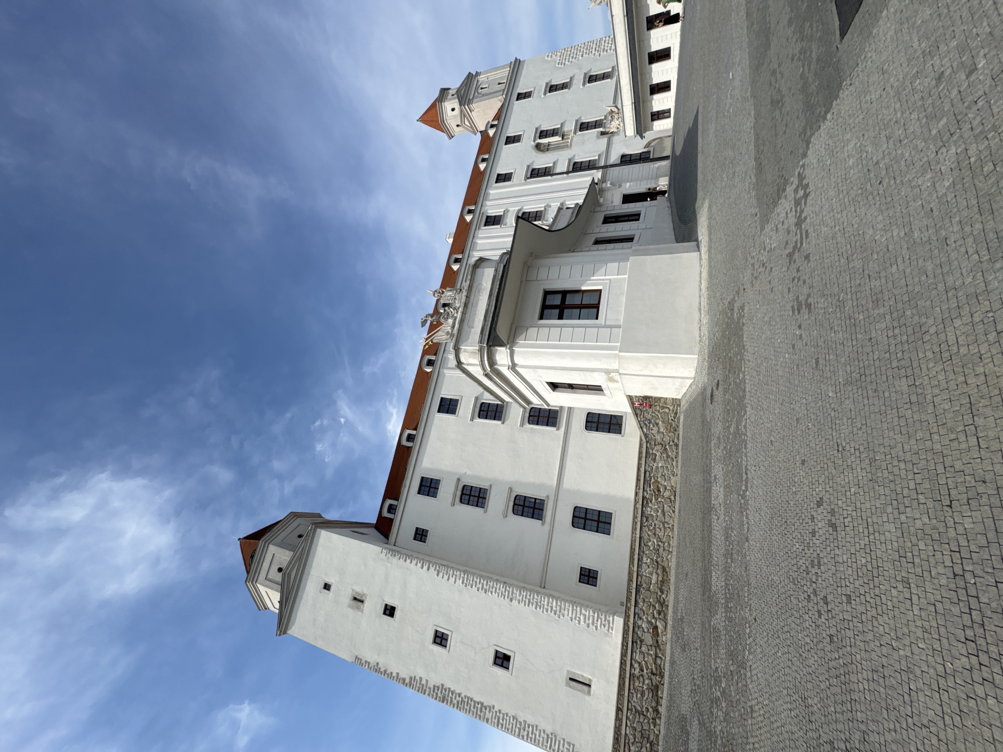 This is a low-angle, vertical photograph of Bratislava Castle in Bratislava, Slovakia. The castle is a large, white building with red-tiled roofs and several prominent towers. The architecture is grand and imposing, with multiple windows and decorative elements. The foreground is paved with cobblestones, and a stone wall runs along the base of the castle. The sky above is a bright blue with scattered white clouds, suggesting it is daytime. The scene is tranquil and hints at a pleasant day for exploring a historical landmark. There are a few people visible on balconies and in the courtyard, casually observing the surroundings. The overall impression is one of historical significance and architectural beauty.