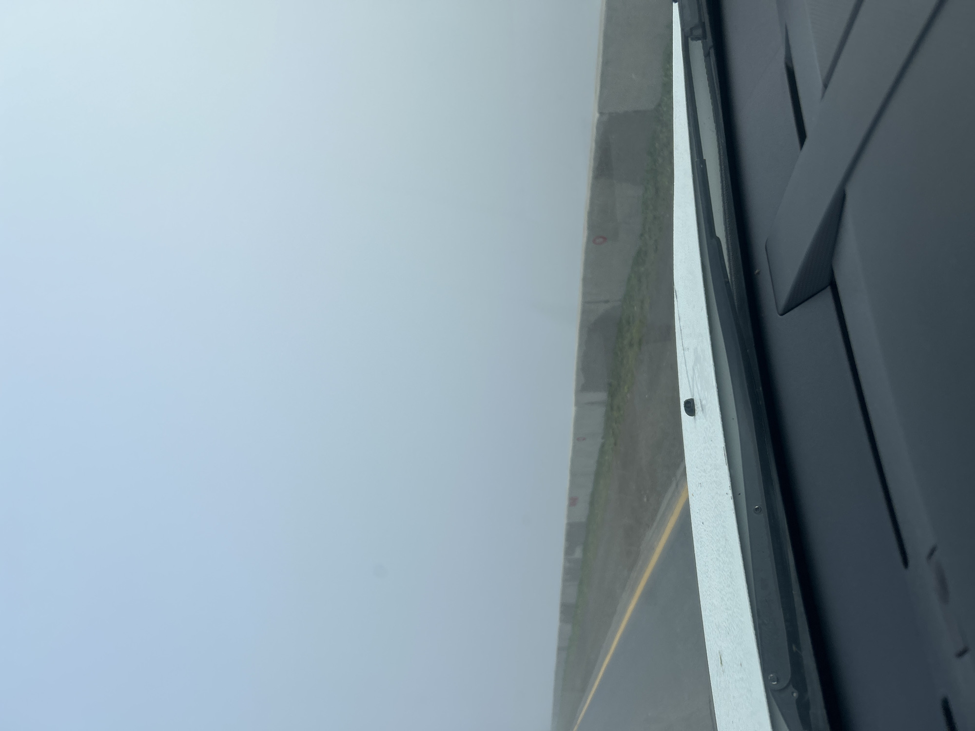 The image is a first-person perspective from inside a vehicle traveling on a road shrouded in thick fog. The road has a yellow line marking the lane. To the right of the road is a concrete barrier with some red circular markings on it. The sky is a uniform, pale blue due to the fog. The foreground shows the dashboard and windshield of the vehicle. There are no people or animals visible. The scene appears to be a highway or a large road outside of a city, given the lack of buildings and the open landscape suggested by the barrier and the expansive, fog-covered horizon. The image does not provide any specific visual cues to definitively place it within Chaikovsky, Russia; it is a generic foggy road scene.
