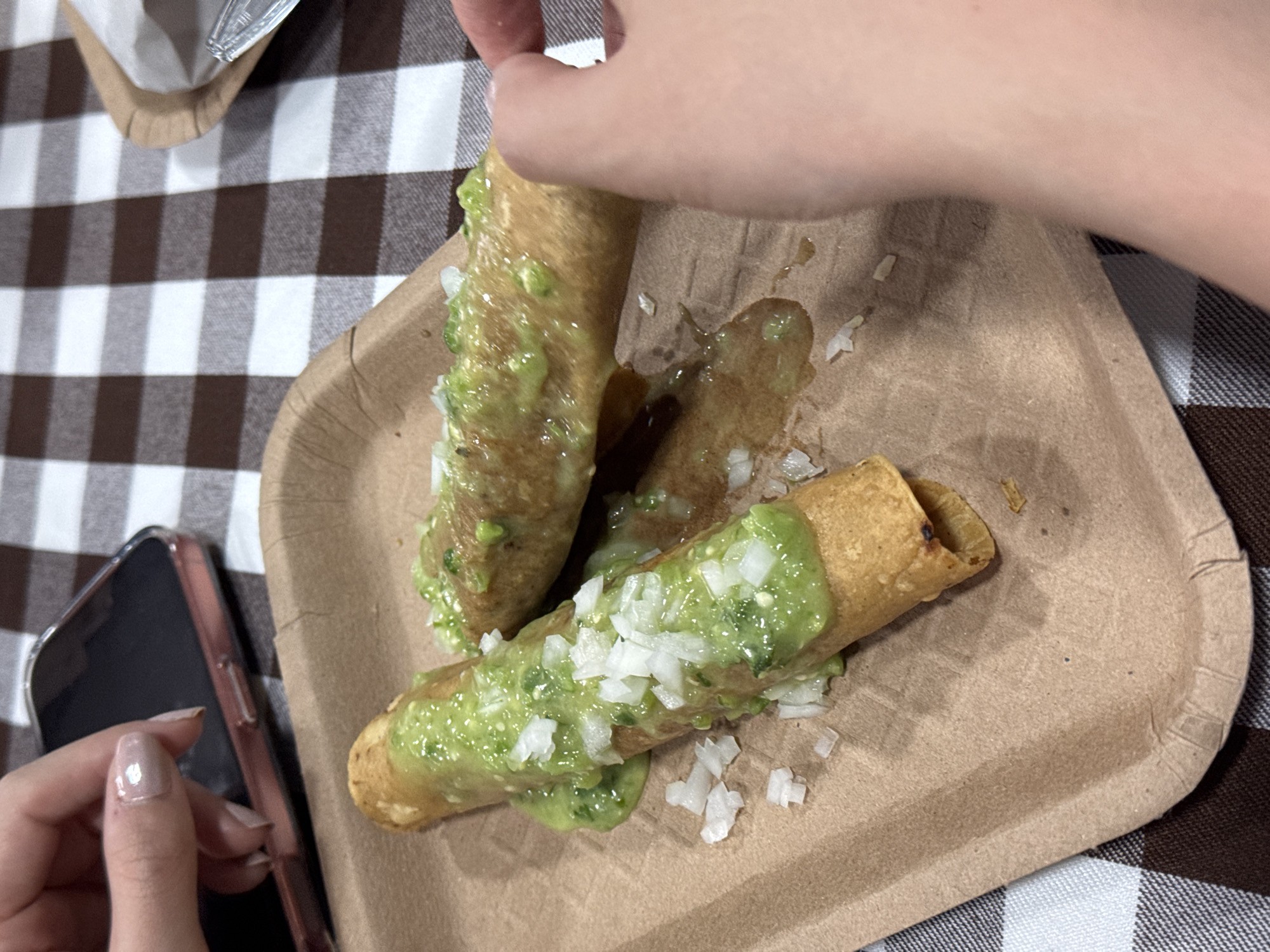 The image depicts a close-up view of a disposable brown paper plate holding three taquitos (rolled tacos) topped with a green sauce, likely guacamole or a similar avocado-based sauce, and diced white onions. A hand is reaching toward the taquitos. A smartphone lies near the plate and a checkered tablecloth is visible in the background. It is not possible to confirm that the location is San Bernardino Tlaxcalancingo, Mexico.