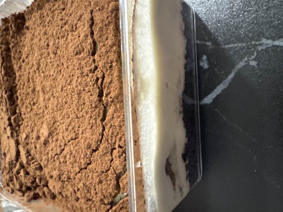 A layered dessert, contained within a clear, rectangular plastic vessel, is depicted resting on a dark, veined surface. The uppermost layer consists of a substantial quantity of dry, brown cocoa powder, exhibiting an uneven, textured, and slightly cracked appearance. Below this, a thick, smooth, off-white creamy layer is visible through the transparent container. The dessert's base comprises a dark, granular layer, likely a crushed biscuit or cake component. The underlying surface is dark gray or black with irregular, light gray or white striations, consistent with a polished stone countertop. No individuals or active events are present in the scene. The location is Rostov-on-Don, Russia.