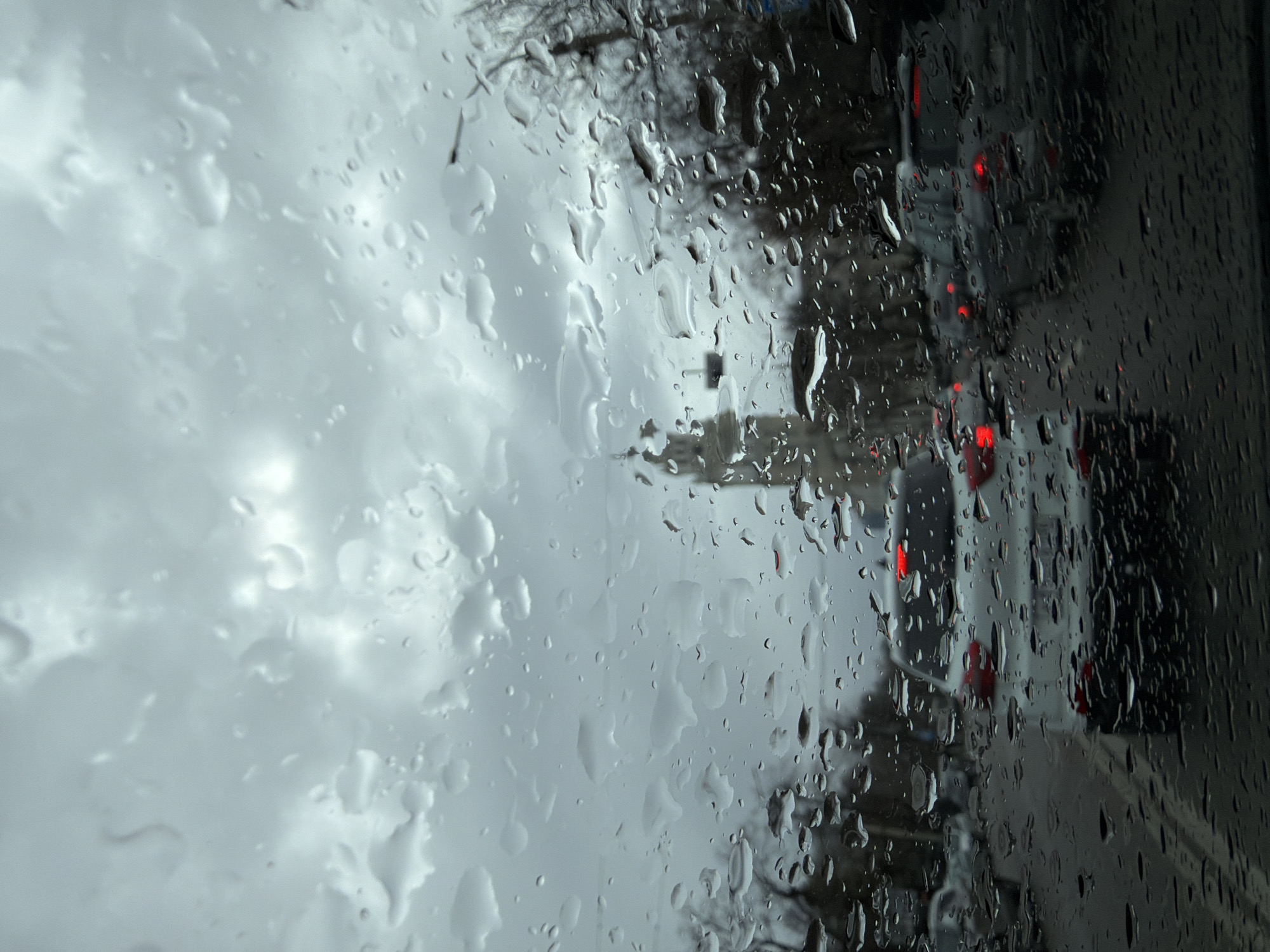 The image is a view through a rain-streaked window. On the left, the distorted view shows a cloudy sky. On the right side of the window, blurred vehicles with red taillights can be seen on a road.