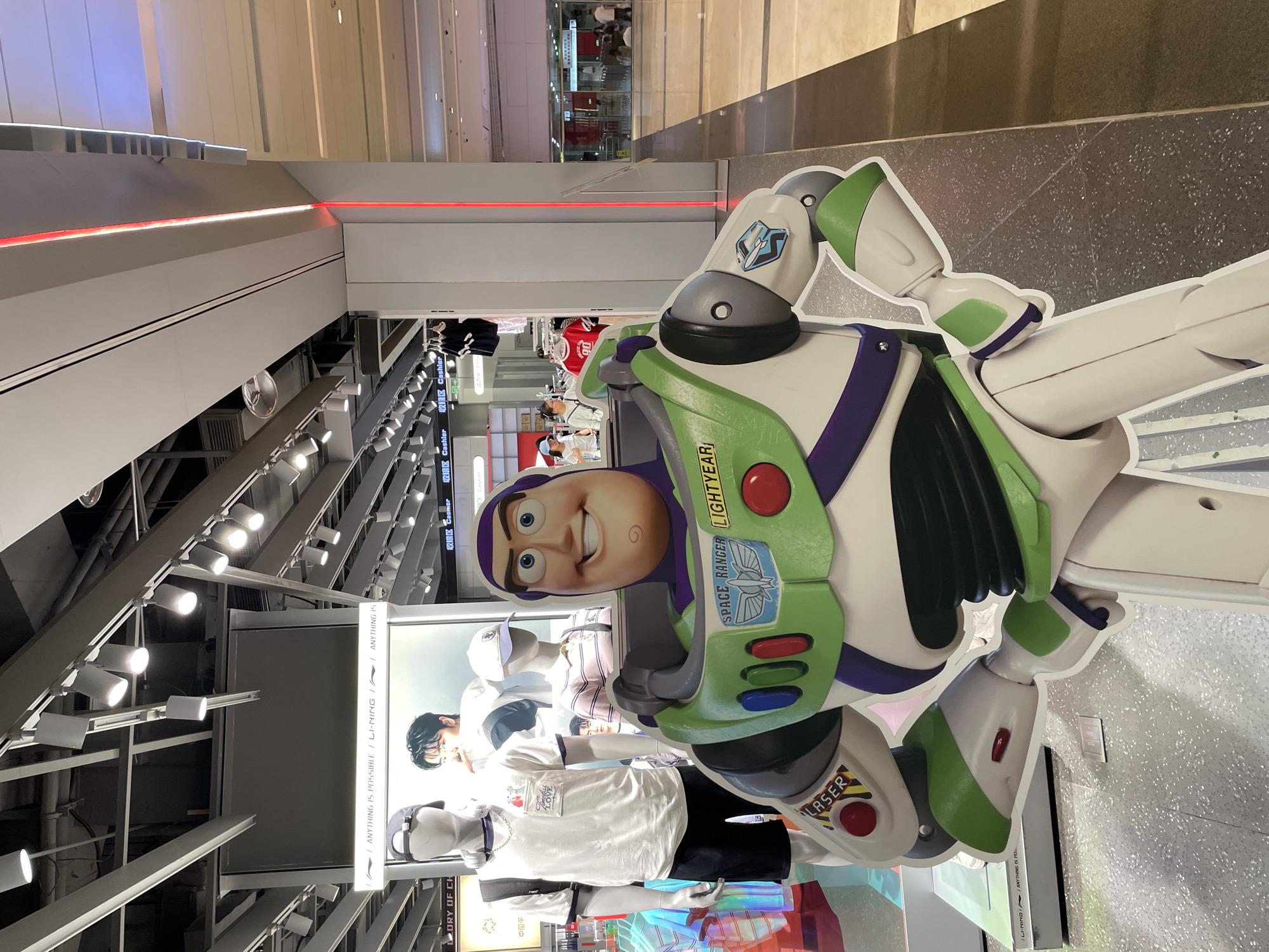 In a retail environment, a life-sized cardboard cutout of Buzz Lightyear from Toy Story stands prominently in the foreground. The cutout is positioned to the right of the frame and faces forward, with its right arm bent and hand on its hip, and its left arm extended slightly. Behind the Buzz Lightyear cutout, mannequins are displayed in athletic apparel, with one mannequin to the left wearing a white t-shirt and black shorts, and another mannequin in the middle wearing a striped pink and white polo shirt. A large advertisement featuring a young man in athletic wear is visible behind the mannequins. The store interior features overhead lighting, clothing racks with apparel, and a floor with a speckled pattern. In the background, through glass doors, a busy mall environment with people can be observed. A red LED light strip runs vertically along a pillar to the right of the Buzz Lightyear cutout. The store appears to be a Li-Ning store, indicated by signage. The setting is within a shopping mall in Yuzhong District, China.