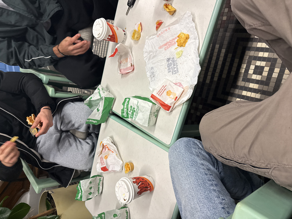 A table covered in fast food wrappers and containers suggests a recent meal. Used cups, crumpled paper bags labeled "Onion Rings," and discarded food containers litter the surface. Two people are seated at the table, one is eating fries and the other is wearing a dark jacket. Another person with blue jeans is seated at the table as well. A patterned floor is visible.