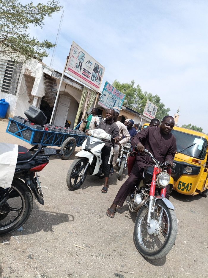 The image depicts a street scene in Gama, Nigeria, under a bright, clear sky. Multiple individuals, predominantly adult males, are present on a dusty or unpaved road. In the immediate foreground, a man wearing a dark brown long-sleeved garment and dark pants sits on a red and chrome motorcycle, looking towards the camera. He wears dark flip-flops. Directly behind him, another man in a dark grey shirt and dark pants is seated on a white and black motorcycle, also facing forward. Several other men on motorcycles are visible further back in the line of vehicles. To the right, a yellow auto-rickshaw (Keke Napep) with the number "6543" on its side is partially visible. To the left, a blue metal two-wheeled trailer, laden with multiple black jerrycans or containers, is parked or being towed. One black container rests precariously on top of the stack.
The background features a dilapidated building with exposed brickwork and a corrugated metal roof on the left, near which a blue barrel or bin is visible. Above this building, a large white billboard sign, advertising what appears to be various products or services, is mounted. A smaller blue sign is positioned below it. Further back, green trees are visible, and in the far background on the right, a light-colored mosque minaret with a pointed top rises against the sky. A tall, slender antenna or mast is visible in the distance on the far left. The scene suggests a busy thoroughfare or commercial area with pedestrian and vehicular traffic.