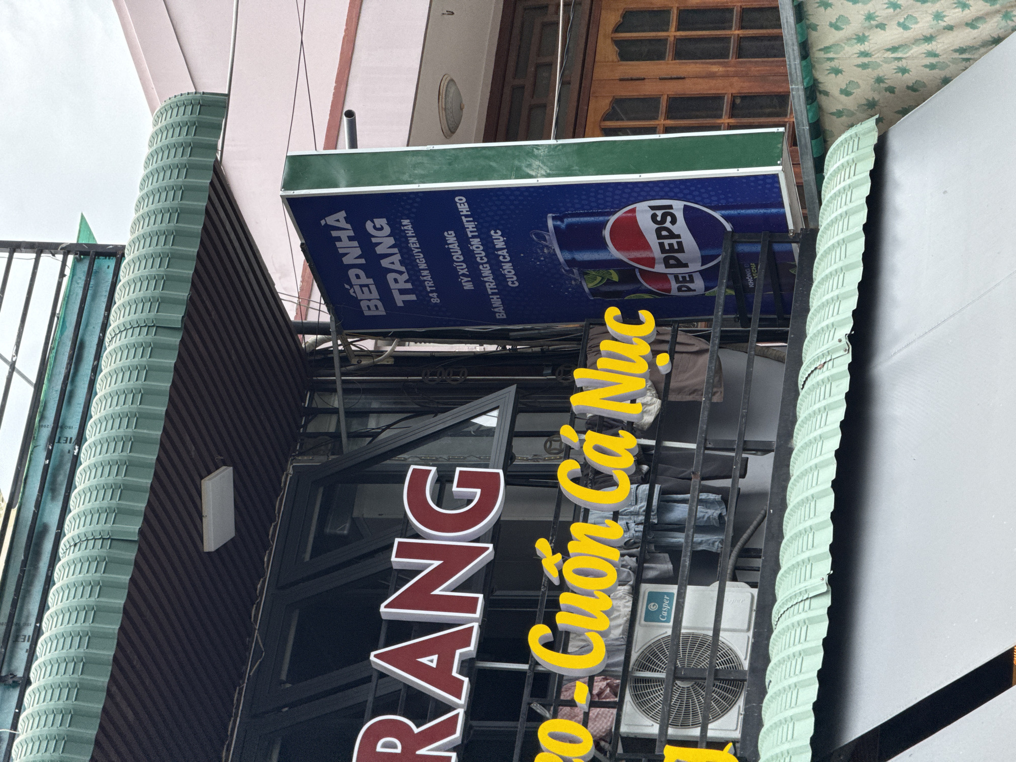 The image depicts a building facade in Nha Trang, Vietnam, featuring prominent signage for a restaurant. A large red and white sign spells out "TRANG" in large block letters. Below this, a yellow, cursive sign reads "Co - Cuốn Cá Nục", indicating a dish of fish rolls. To the right, a vertical blue sign with green trim displays text in Vietnamese, including "BẾP NHÀ TRANG" (Nha Trang Kitchen) and an address "84 TRẦN NGUYỄN HÁN". The sign also features images of Pepsi cans and lists food items such as "MỲ XỬ QUẢNG", "BÁNH TRÁNG CUỐN THỊT HEO", and "CUỐN CÁ NỤC". An air conditioning unit is visible on a balcony-like structure in front of the building. The overall setting appears to be an urban environment, with awnings and neighboring buildings visible in the background. The lighting suggests it is daytime.
