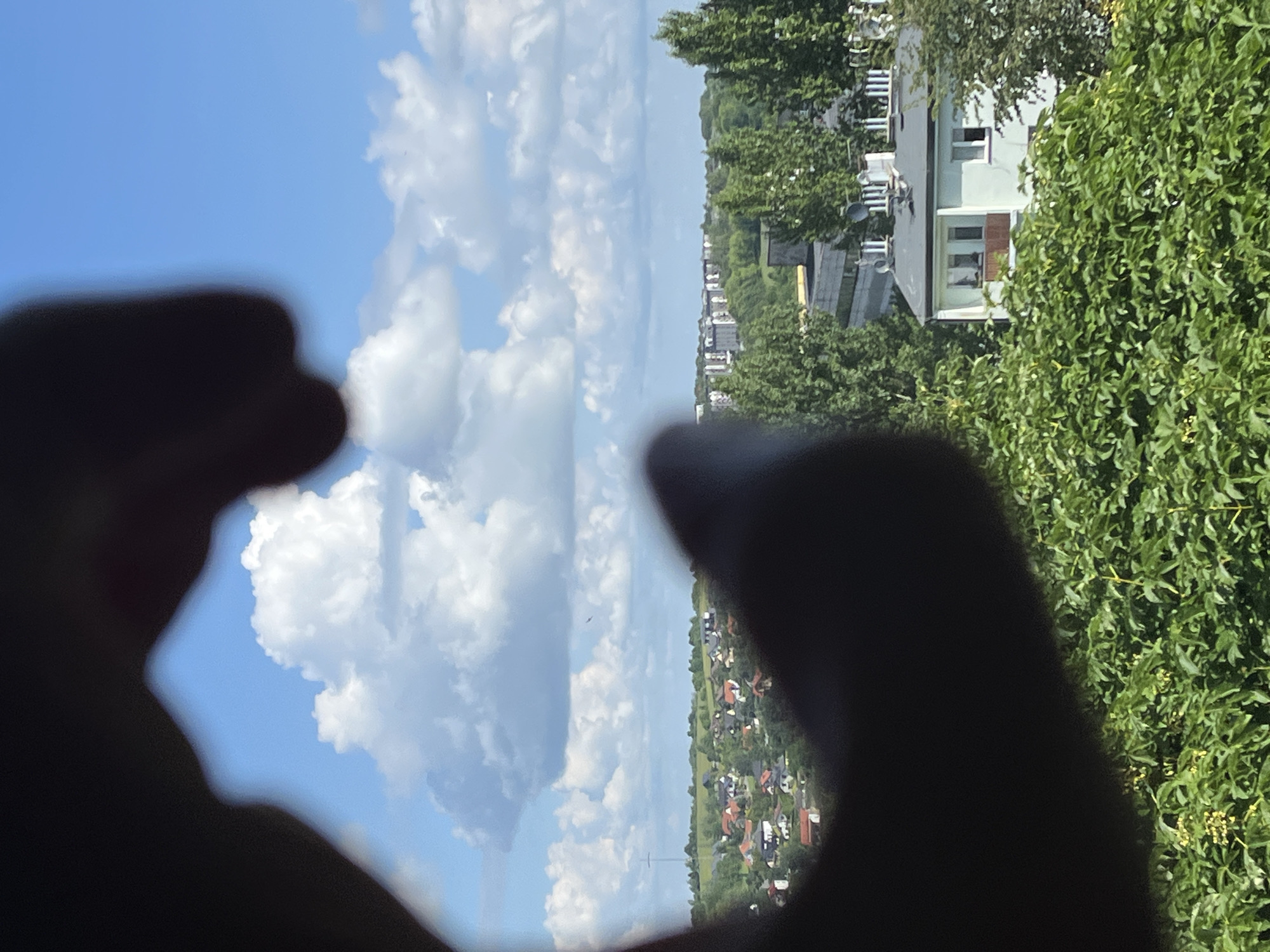 A hand is positioned in the foreground, partially obscuring the view of a distant cityscape. The hand is dark and silhouetted against a bright blue sky filled with white, puffy clouds. Through the opening created by the hand, a village or small town can be seen with red-roofed houses nestled among green trees. In the mid-ground, a cluster of apartment buildings is visible, and further in the distance, taller buildings suggest a more urban area. The scene is set on a sunny day, with lush green foliage in the lower portion of the image.