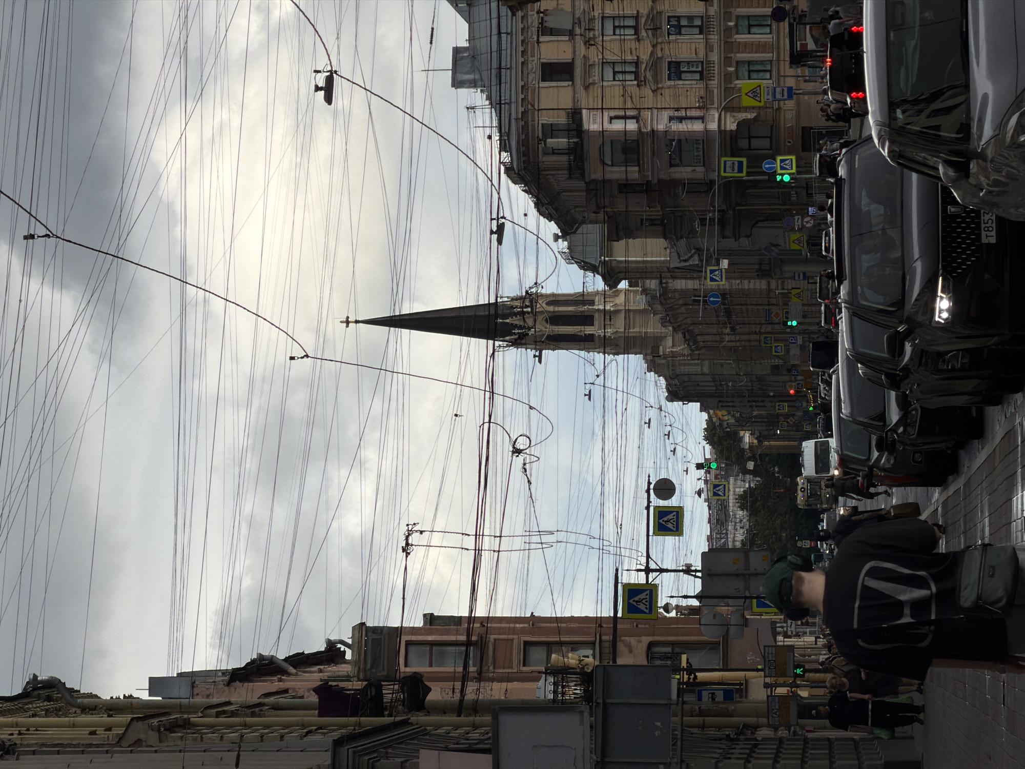 This is an eye-level, medium shot of a street in Saint Petersburg, Russia, looking down a wide boulevard lined with multi-story buildings. The sky is overcast with a dense layer of gray clouds, and a complex network of overhead wires crisscrosses the frame. The architecture is predominantly European, with ornate facades and balconies visible on the buildings.
In the foreground, a young man wearing a dark t-shirt with a white logo, a green baseball cap, and headphones is walking away from the camera. He has a dark green crossbody bag slung over his shoulder. He is walking on the sidewalk, which is lined with parked cars to his right. Several other pedestrians are visible on the sidewalk, some walking towards the camera and others walking in the same direction as the foreground subject.
The street itself is busy with vehicular traffic. Several cars are stopped at what appears to be an intersection, indicated by traffic lights and numerous road signs, including pedestrian crossing signs. A white van or bus is visible further down the street. In the distance, a tall church spire with a cross on top pierces the cloudy sky, a prominent landmark in the city. The overall atmosphere of the scene suggests a typical urban day in Saint Petersburg, with cloudy weather and moderate pedestrian and vehicular activity.