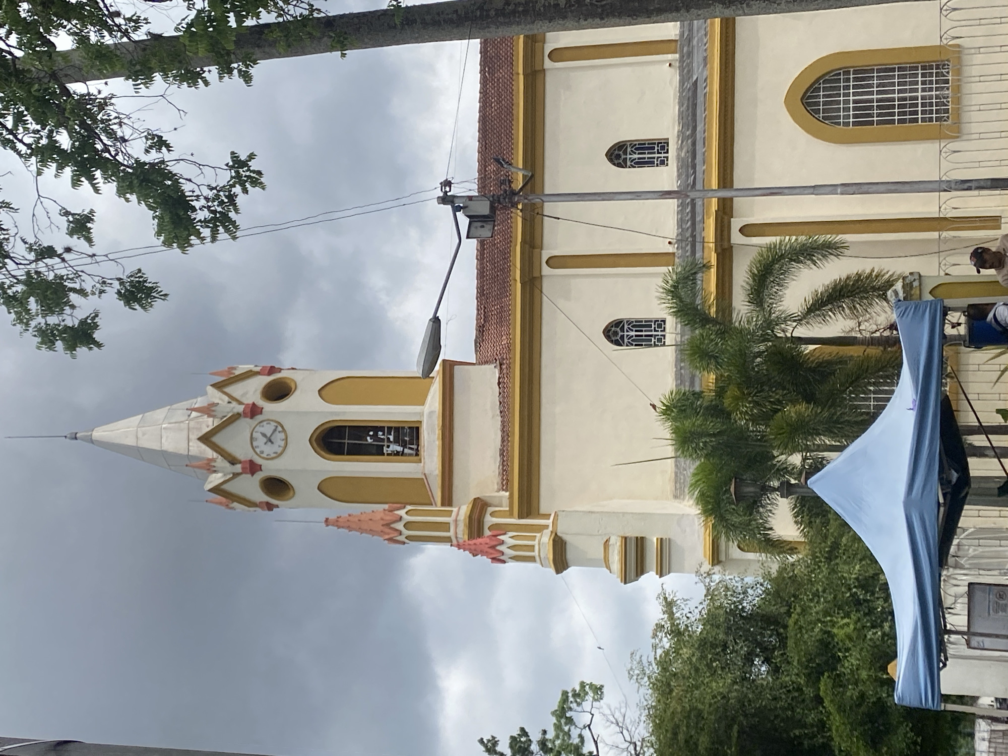 The image shows a church building in Tinaquillo, Venezuela. The church has a tall, pointed tower with a clock and steeple. The building is cream-colored with yellow and pink trim. The sky is cloudy. A blue canopy is partially visible on the right side of the image, and there are trees in the foreground. A light pole with wires is visible in front of the building.
