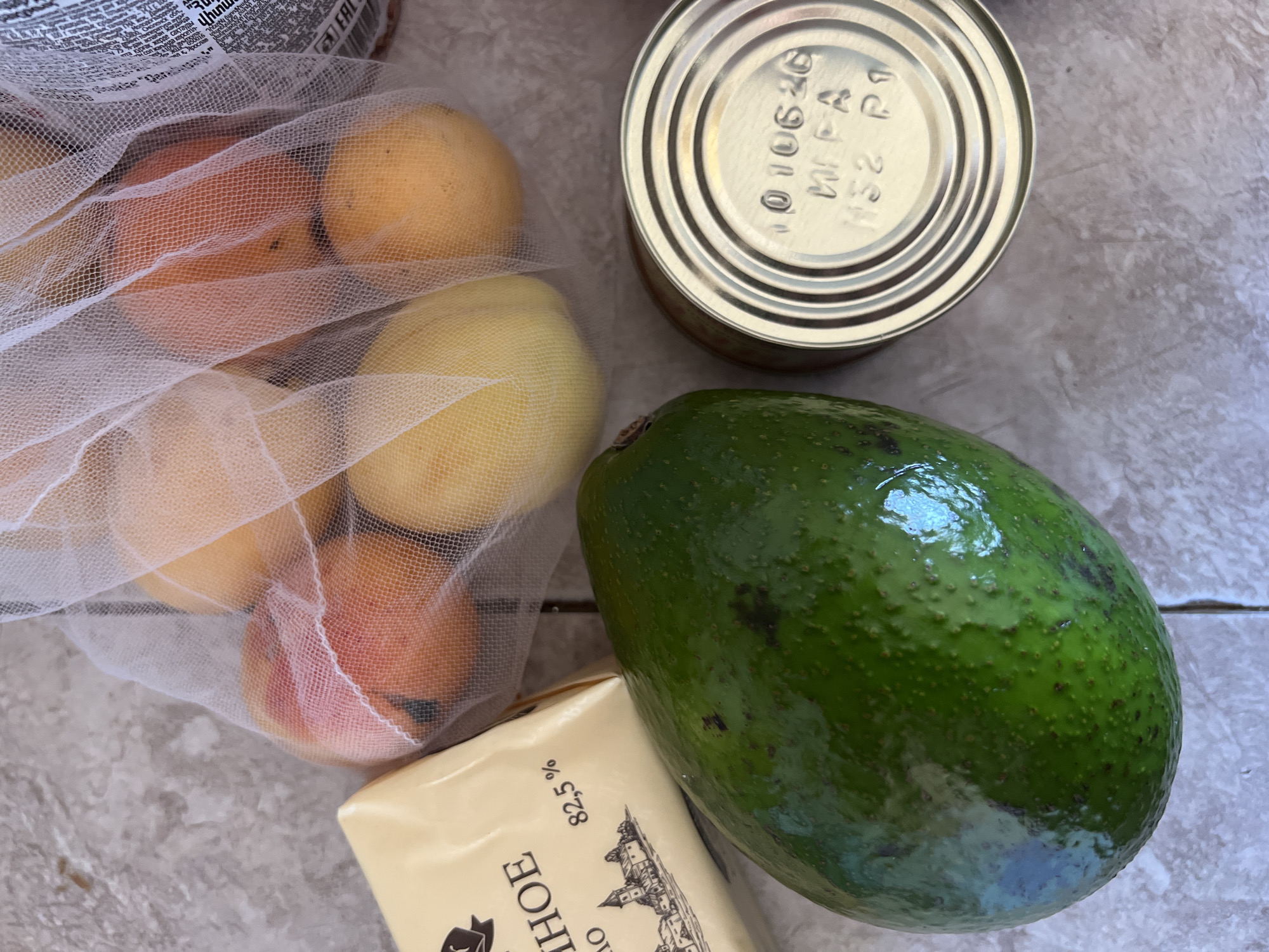 The image shows a close-up, overhead view of groceries on a tiled surface. The items include an avocado, a block of cheese, a mesh bag containing several apricots, and a tin can. The cheese packaging is yellow and displays text in Cyrillic and "82.5%". The tin can has a stamped expiration date of "010620" and other alphanumeric characters. The background is a light-colored, speckled tile. There is no indication of the city of Novosibirsk or any other specific location.