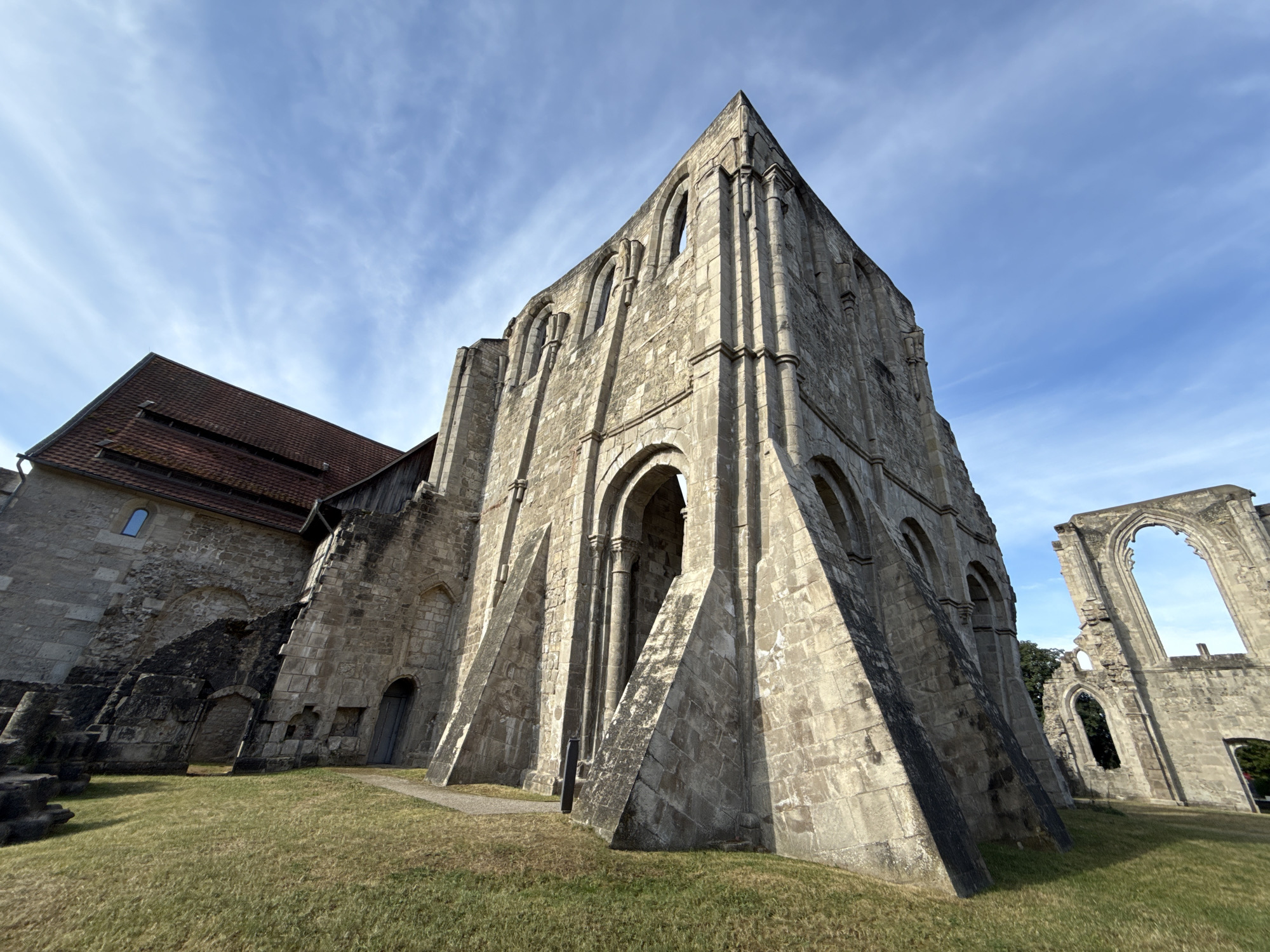 The image depicts a section of the ruins of Walkenried Abbey, a Cistercian monastery located in Walkenried, Germany. The primary focus is on a large, weathered stone structure, likely a former church or abbey building, characterized by its Gothic architecture. Arched windows, some intact and others open to the sky, are visible along the crumbling facade. The stone is a light gray, showing signs of age and erosion. The structure is partially intact, with large sections missing, revealing the skeletal remains of the building.
To the left of the main ruin, a portion of a building with a red-tiled roof is visible. This section appears more preserved, with a single small, rectangular window visible in a stone wall. The ground in front of the ruins is covered in dry, yellowish-green grass. A paved pathway, light gray in color, leads towards the entrance of the main ruin.
In the background, to the right of the main structure, another section of ruin is visible, featuring a large, arched opening against the blue sky. The sky itself is bright blue with thin, wispy white clouds. The overall impression is one of historical grandeur in a state of decay, set against a clear, sunny day. There are no people visible in the image. The scene is static and appears to be a daytime shot, with the sun casting shadows that highlight the texture of the stone and the contours of the ruins.