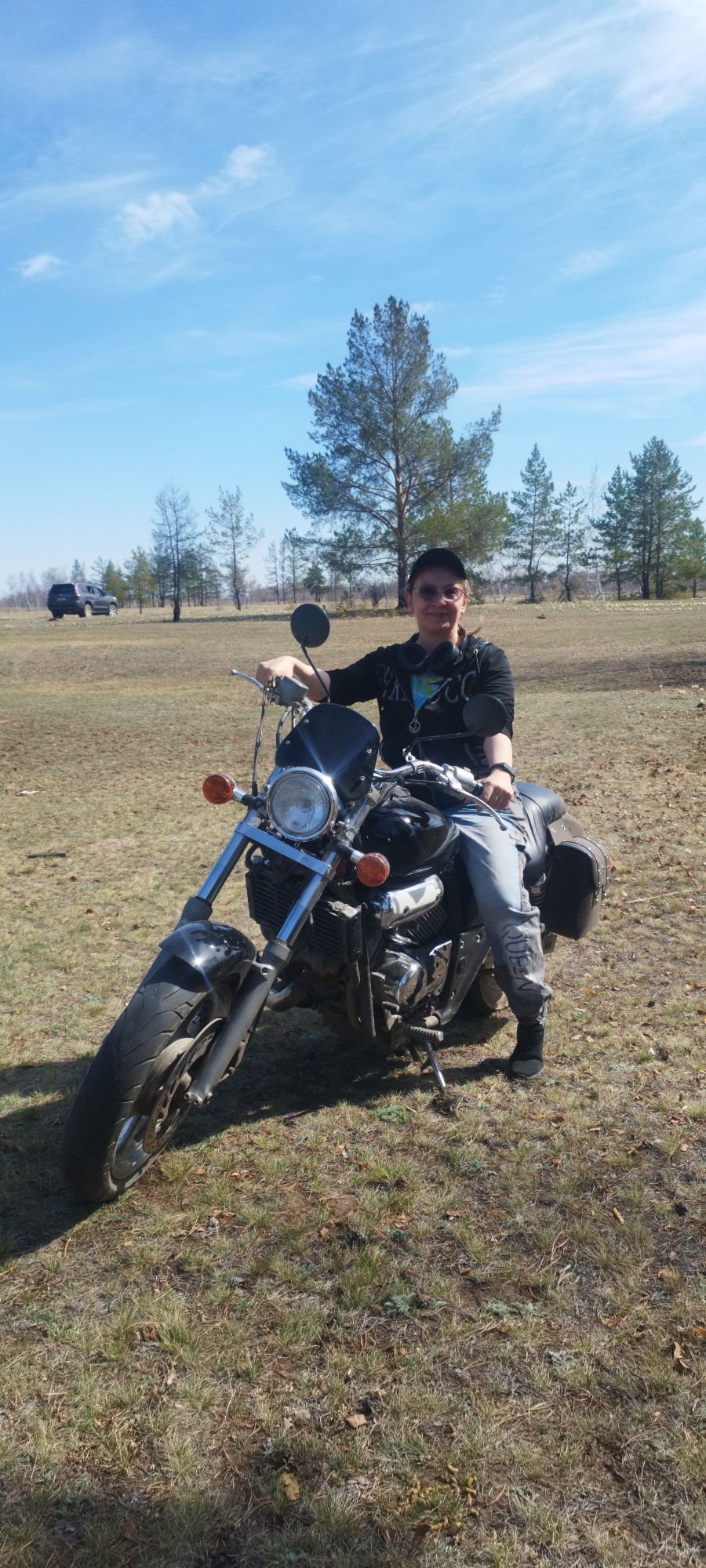 A person wearing a dark jacket and light-colored pants sits on a black motorcycle in a grassy field. The person also wears a dark cap, glasses, and a necklace. The motorcycle has a front windshield, headlight, and a leather saddlebag. Behind the motorcycle and person, there is a field with scattered trees under a blue sky with light clouds. A dark SUV is parked in the distance. The location appears to be a rural area near the city of Syrdakh, Russia.