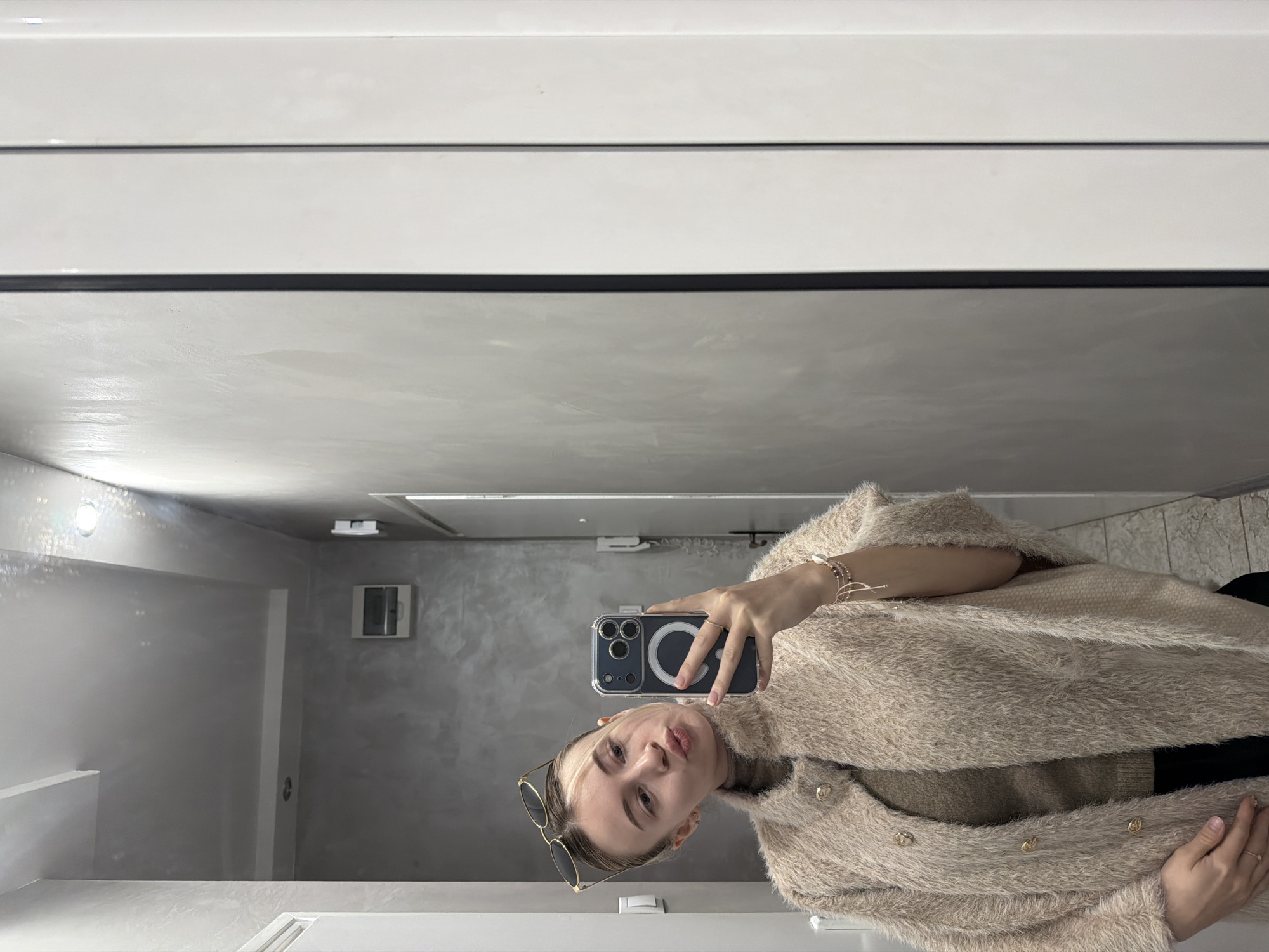 A woman is taking a mirror selfie in a modern bathroom. The bathroom features light gray walls, a recessed light, and a white vanity. The woman is wearing a fuzzy, light brown jacket with gold buttons. She is holding a smartphone with a clear case and a ring holder on the back. Sunglasses are perched on her head. It's impossible to determine if the bathroom is in Plovdiv, Bulgaria, as there are no identifying details within the image that specify location.
