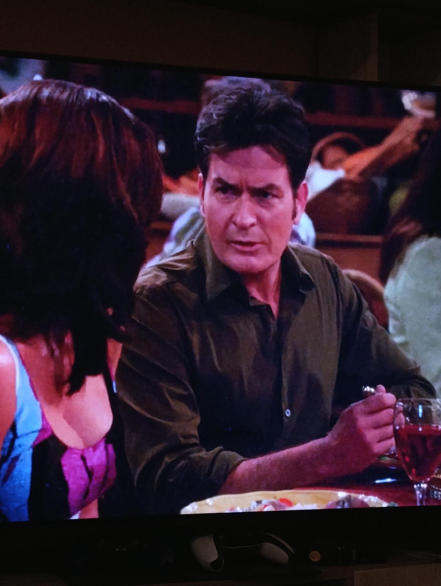 The image shows a scene from the TV show "Two and a Half Men". Actor Charlie Sheen, in character as Charlie Harper, is sitting at a table in what appears to be a restaurant. He is wearing a dark green button-down shirt and holding a fork in his right hand. A glass of red wine sits on the table next to a plate of food. Opposite him sits a woman with dark hair, wearing a blue and purple top. The background is blurry, suggesting a busy restaurant environment with other patrons. This image does not appear to be in Plovdiv, Bulgaria; "Two and a Half Men" was filmed in California, USA.