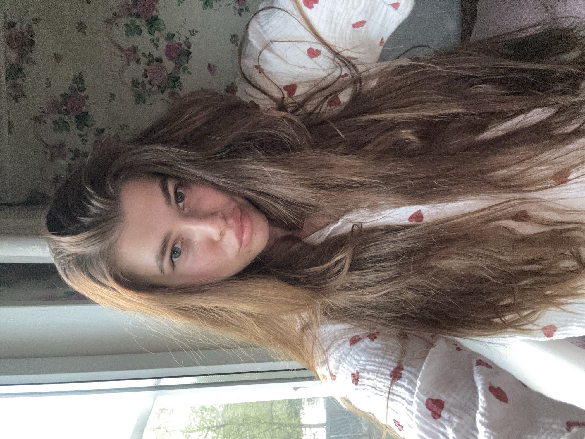 A young woman is positioned near a window, her face visible in the frame. She has long, light brown hair. She is wearing a white garment with red heart patterns. The window looks out onto greenery. The background includes floral wallpaper. There is no information to support that this image was taken in the city of Мещёры, Russia.