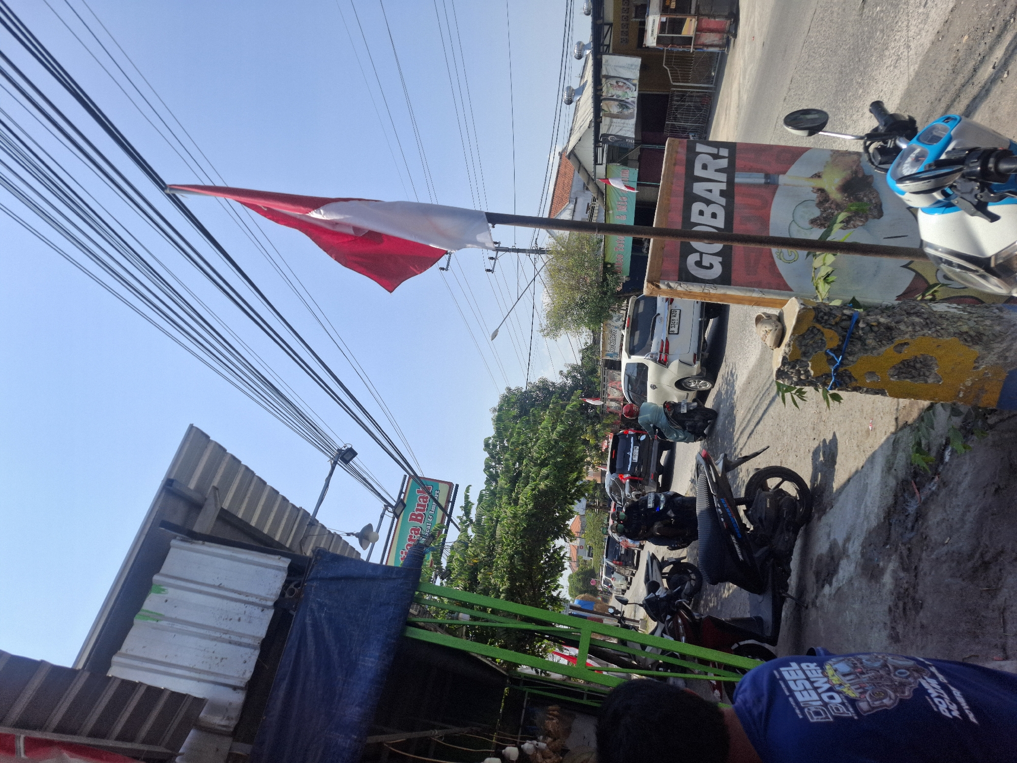 In Sumberwudi, Indonesia, under a clear blue sky, a street scene unfolds. The foreground features a person viewed from the back, wearing a blue t-shirt with the text "DIESEL POWER." Adjacent to them, a row of parked motorcycles, including a black scooter and a red scooter, line a sidewalk. The sidewalk is weathered and cracked, with a post adorned with yellow and blue paint. A sign with the word "GOBAR!" is prominently displayed, featuring an image of food.
Beyond the sidewalk, a street is congested with traffic, including cars and motorcycles, indicating a busy urban environment. A red and white flag, likely the Indonesian flag, waves from a pole. The background shows buildings with corrugated roofs, trees, and overhead power lines. A sign with green lettering that appears to say "Niaga Buah" (Fruit Trade) is visible on a building. The overall impression is a bustling street in a part of Indonesia, possibly during a festive or busy period, given the presence of the flag and the "GOBAR!" sign, which could be an advertisement or an announcement.