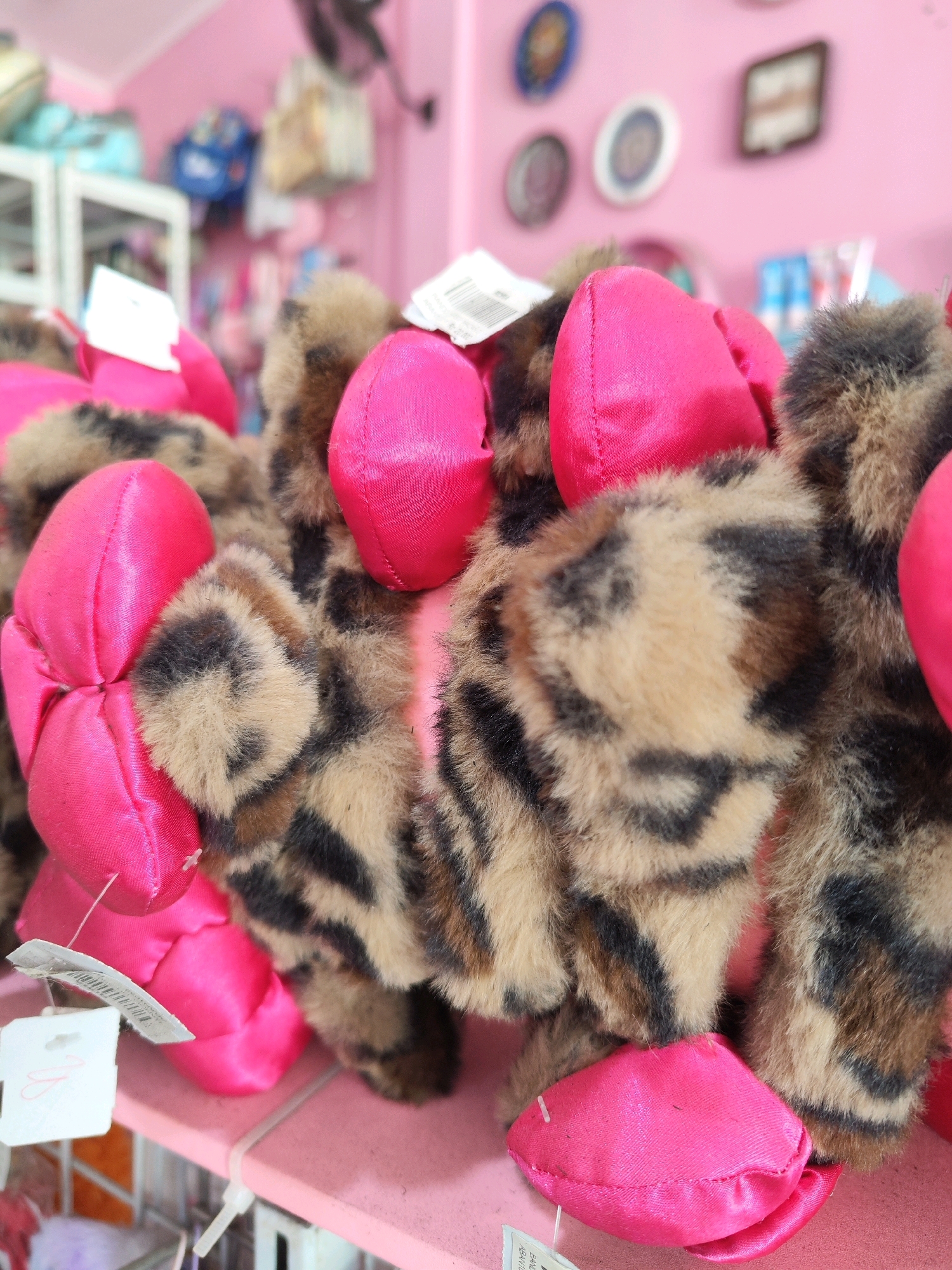 A close-up, eye-level shot shows a stuffed toy with leopard print fur and bright pink accents. The toy appears to be a cat or a similar animal, with its paws and body adorned with the pink material, possibly forming paws or decorative elements. The toy has tags attached, indicating it is new and for sale. The background is blurred but suggests a retail environment, with shelves, other toys, and pink walls. The overall scene is brightly lit and evokes a playful atmosphere.
This image was taken in the city of Sragen, Indonesia. The specific location is not discernible beyond being a retail setting.