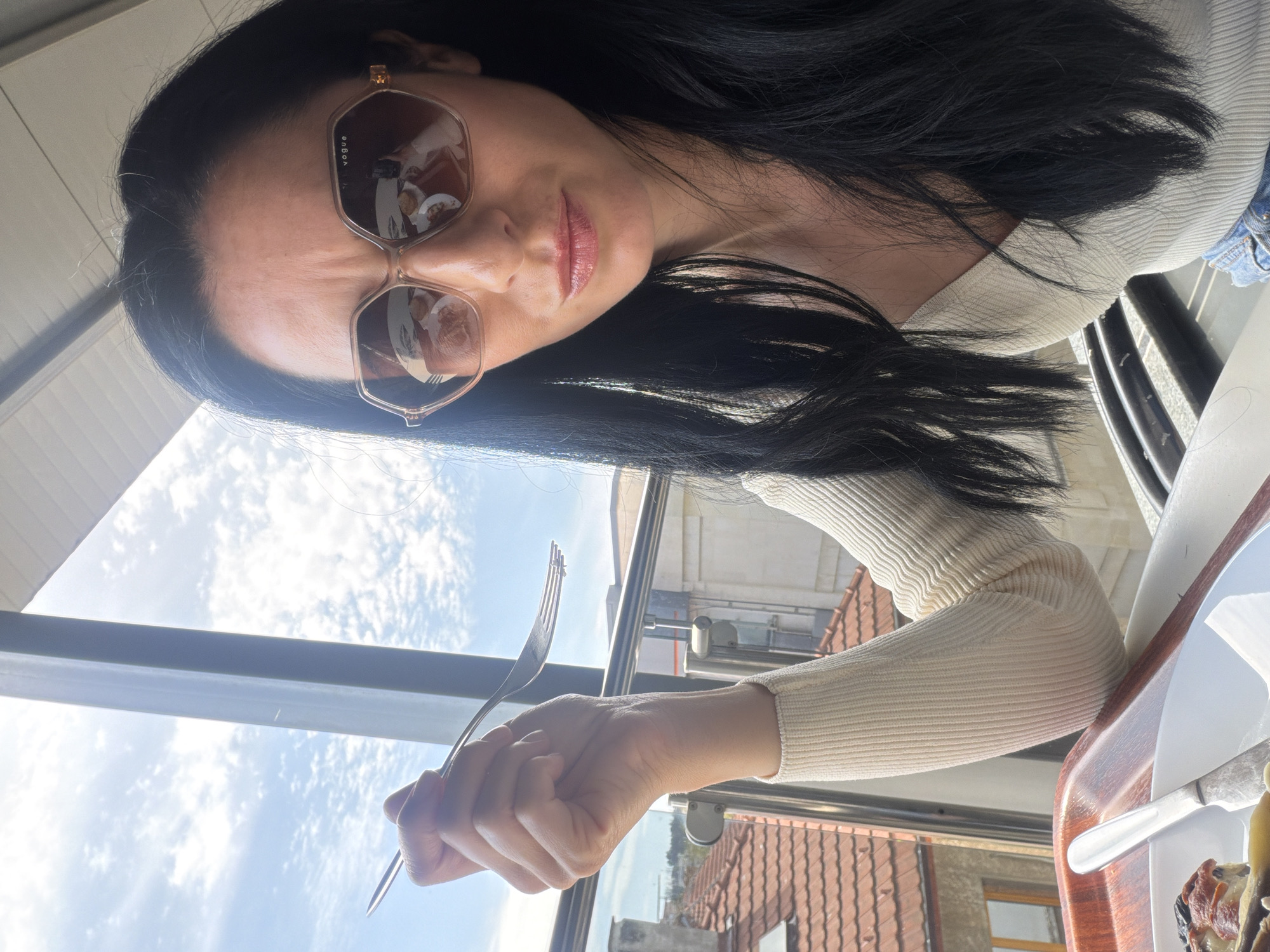 The image is a close-up, angled selfie featuring a woman indoors, looking towards the camera. She has long, dark hair and is wearing large, hexagonal-shaped sunglasses with "VOGUE" text visible on the right lens. Her lips are a glossy light pink. She is dressed in a cream or off-white ribbed long-sleeved top, with a glimpse of blue denim fabric near her left shoulder.
In her right hand, she holds a silver fork, positioned against the bright sky visible through a window. To her right, a partially visible white plate with food (appearing to be a baked item or savory dish with toppings) rests on a wooden surface.
Through the large window behind her, a clear blue sky with scattered white clouds indicates a sunny daytime. Below the sky, traditional red-tiled rooftops and the upper parts of other buildings are visible, suggesting an urban or town setting. The reflections in her sunglasses show indistinct shapes. This scene is set in Pleven, Bulgaria.
