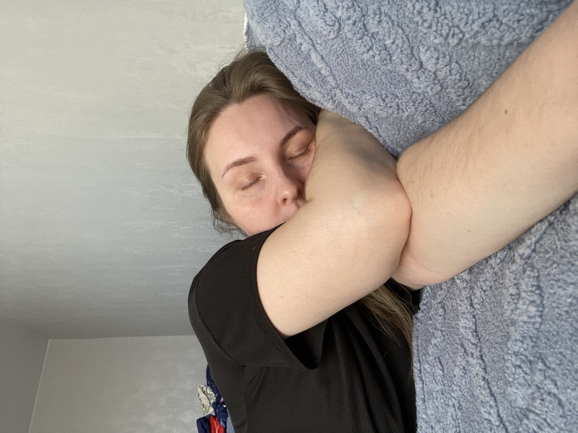 A person is lying with their face pressed against a plush, light blue object, likely a blanket or pillow. The person has fair skin, closed eyes, and light brown hair. They are wearing a black shirt. The background consists of a plain white wall and a glimpse of possibly a curtain or clothing rack in the lower-left corner. The location is not discernible from this image.