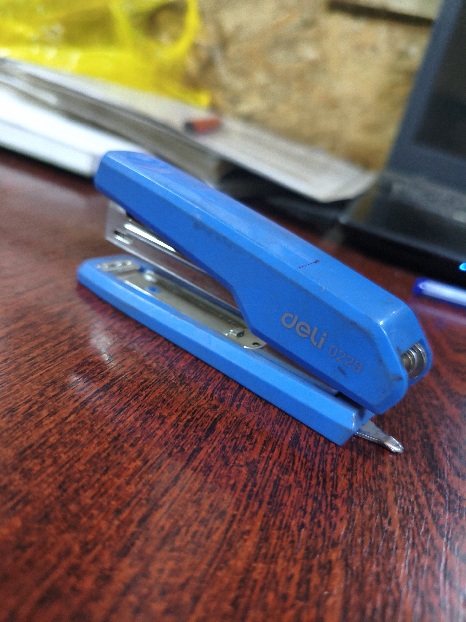 A blue stapler is placed on a wooden surface. The stapler has the brand name "deli" and the model number "0229" printed on it. The background shows out-of-focus objects, including what appears to be papers or a document and a black electronic device. The lighting suggests an indoor environment. There are no people visible in the image. The specific location of Termirtaw city administration, Kazakhstan is not depicted or identifiable from the provided image.