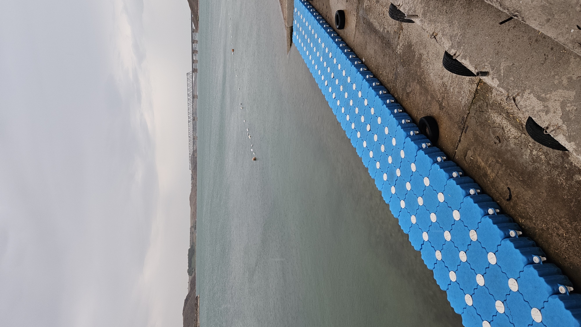 An outdoor, eye-level, vertical shot captures a scene of a long, blue, modular floating dock extending from a concrete embankment into a large body of water under an overcast sky. The dock is composed of interconnected hexagonal plastic modules with white circular accents. To the right of the dock, resting on the concrete embankment, are two black tires. Further up the embankment, a series of vertical metal poles with more black tires attached at different heights are visible, suggesting a form of barrier or erosion control. The water is a muted teal color and appears calm. In the distance, across the water, a long, metal truss bridge spans the horizon, with rolling, arid hills visible beyond it. The sky is a uniform, pale gray, indicating a cloudy day. The overall scene is serene and devoid of human presence, with a focus on the man-made structures and the natural landscape of the city of Qonaev, Kazakhstan.