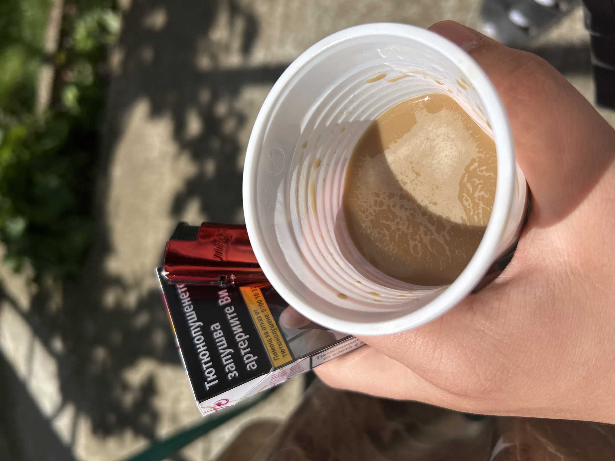 A high-angle shot depicts a hand holding a white, ribbed, plastic cup filled with a light brown liquid, presumably coffee or tea with milk. A pack of cigarettes rests against the cup, with the warning "Тютюнопушенето запушва артериите Ви" (Smoking clogs your arteries) visible, along with a hotline number for quitting smoking. The background is blurry, but suggests an outdoor setting, possibly a yard or sidewalk. Based on the text on the cigarette pack, the image is likely taken in Bulgaria, specifically Koniovets, Bulgaria.