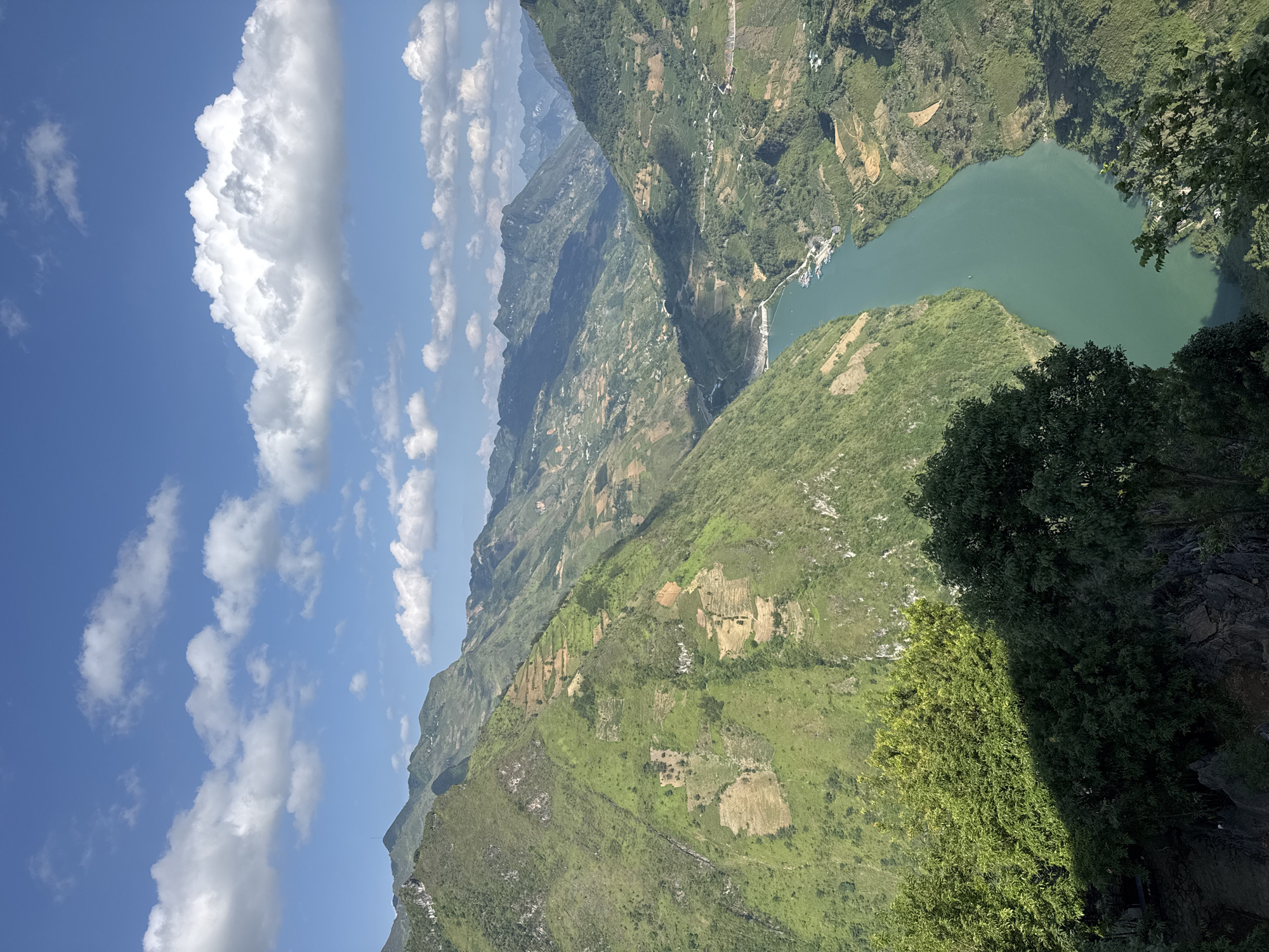An eye-level, rotated image of a mountain range in Đồng Văn Commune, Vietnam, shows a winding river between grassy hills and a blue sky with clouds. The winding river on the right is light green and has docks or boats along its banks. The foreground is filled with grassy hills covered in brush. In the background, there are multiple mountain ranges that fade into the distance. The blue sky in the upper left corner is partially covered with white clouds. A tree is on the right-hand side of the image.