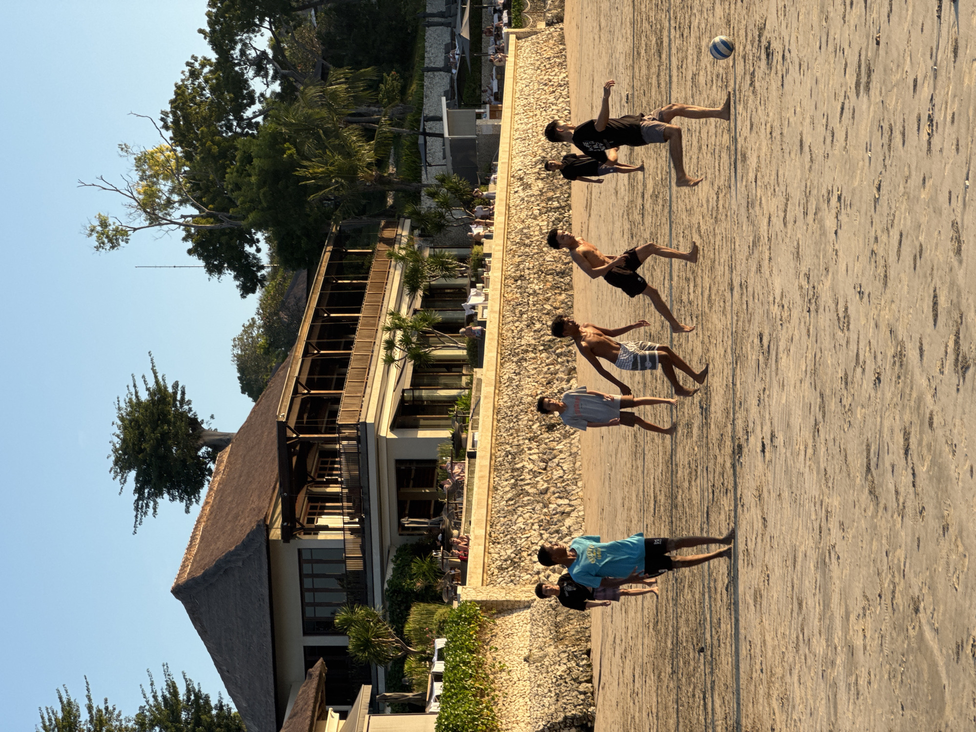A group of young males are playing soccer on a sandy beach in front of a multi-story resort building. The building has a thatched roof and balconies overlooking the beach. The sky is clear and blue, suggesting it is daytime. The people are wearing casual clothing such as t-shirts and shorts. The sand is light brown and appears dry. In the foreground, the feet of the players are visible as they run and kick the soccer ball. The environment is a tropical beach setting with palm trees and lush vegetation surrounding the resort.