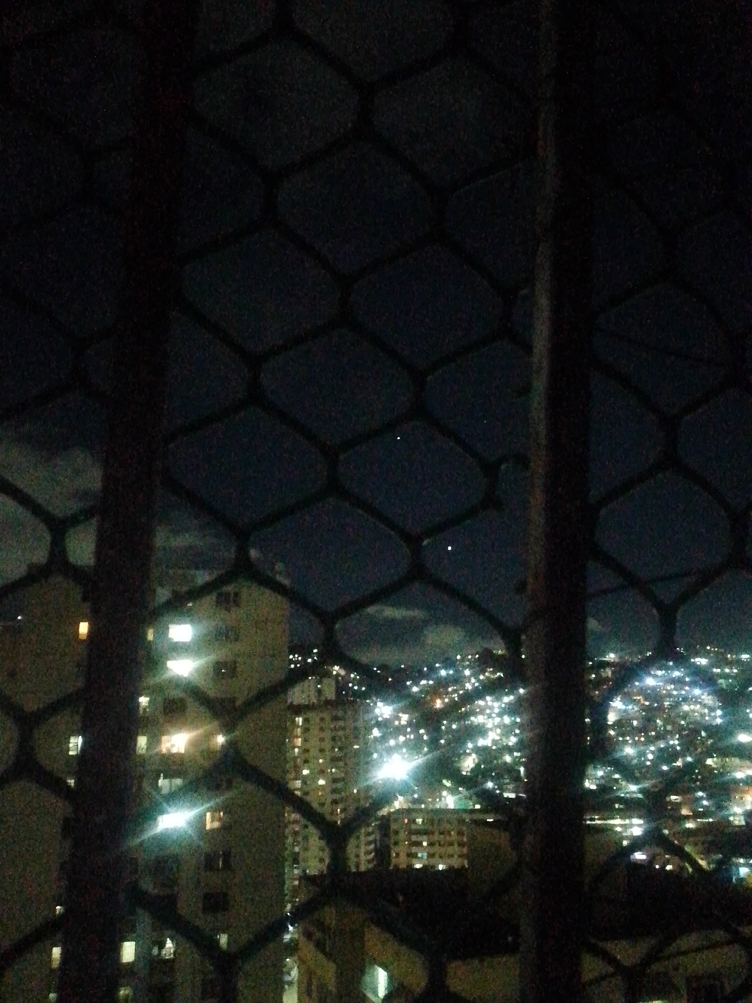 The image shows a night view of Caracas, Venezuela, seen through a metal window grille. The background is a cityscape with illuminated buildings and streetlights spread across hills. There are at least two high-rise buildings with multiple illuminated windows. Two bright dots are visible higher in the sky. The foreground consists of a dark, diamond-patterned metal grille, partially obscuring the view. The image suggests an urban residential setting at night.
