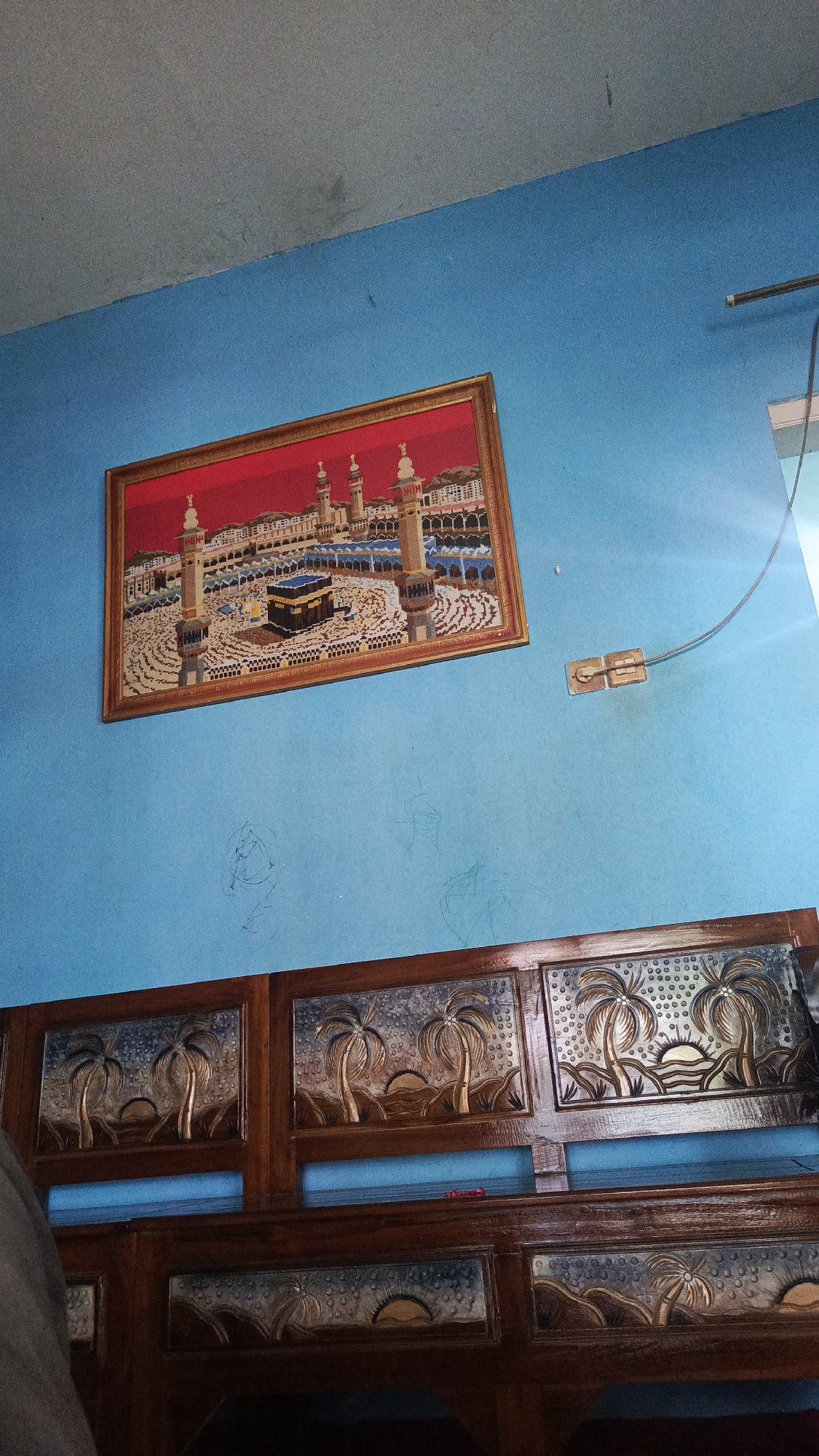 This image captures an indoor scene in a room located in Kajeksan, Indonesia, featuring a brightly painted blue wall, a gray ceiling, and wooden furniture. A rectangular framed artwork depicting the Kaaba and the Grand Mosque in Mecca is hung on the upper left portion of the blue wall, slightly tilted to the right. The artwork appears to be an embroidery or cross-stitch piece with a red background, intricate details of the mosque's architecture, minarets, and the black Kaaba cube, all enclosed within a golden-brown frame.
Below the artwork, the blue wall exhibits several faint, scribbled marks. To the right of the artwork and slightly lower, a beige-colored, surface-mounted electrical outlet with two receptacles is affixed to the wall. A dark-colored electrical wire extends from this outlet upwards and to the right, disappearing out of the frame.
The lower half of the image is dominated by a dark wooden furniture piece, possibly a headboard or a cabinet, which features multiple rectangular panels. The visible panels are adorned with carved or inlaid designs depicting a scenic motif: two palm trees, a rising or setting sun behind rolling hills, and a textured pattern resembling droplets or stars in the sky. At least two full upper panels and partial views of lower panels with similar designs are visible, showing a consistent aesthetic across the furniture. No individuals are present in the scene.