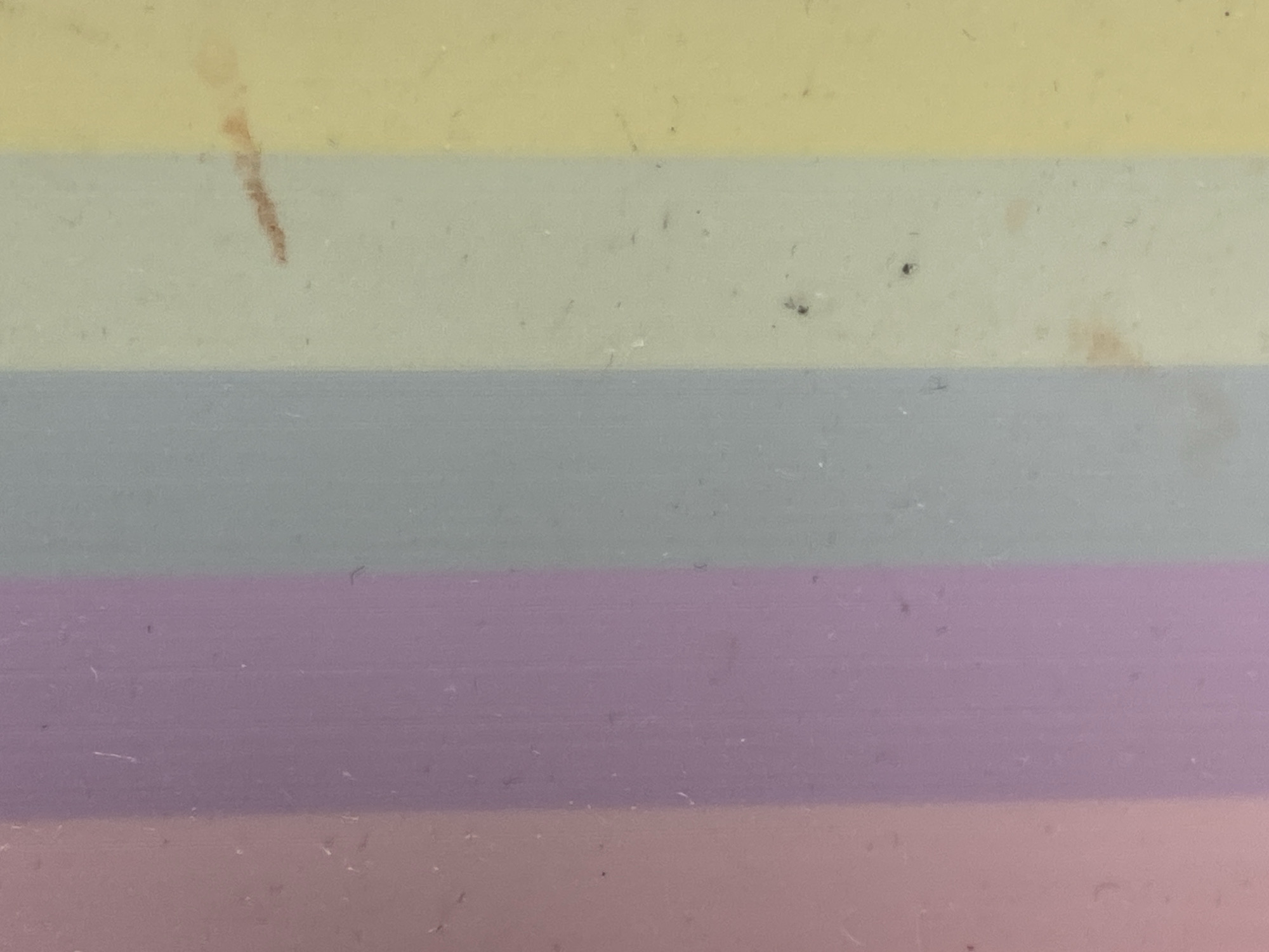 The image displays a close-up view of horizontal bands of pastel colors. From top to bottom, the colors appear to be a muted pink, a light lavender, a pale gray, a pale yellow-green, and a slightly darker yellow. The surface exhibits subtle texture and some minor specks or imperfections. There are no discernible features that would indicate a specific location within Warsaw, Poland, or any other city. The image is abstract and focuses solely on the color and texture of the material.
