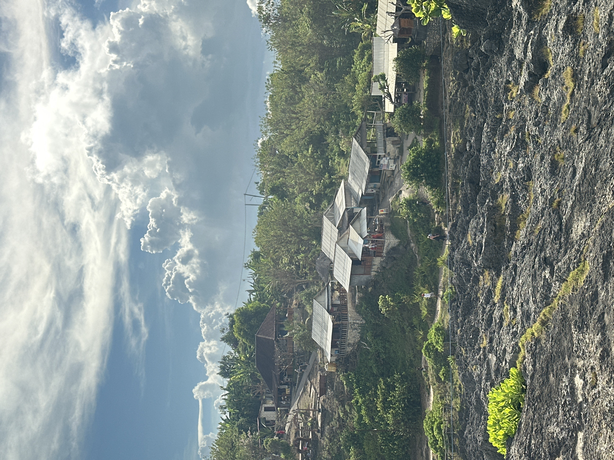The image depicts a view of structures built on a cliffside in Nusa Penida, Indonesia, under a partly cloudy sky. The structures appear to be a mix of traditional and modern designs, possibly residences or businesses. The cliff face is rocky and covered in vegetation. A path runs along the cliff edge, where a few people can be seen walking. The sky is predominantly blue with large, puffy white clouds.