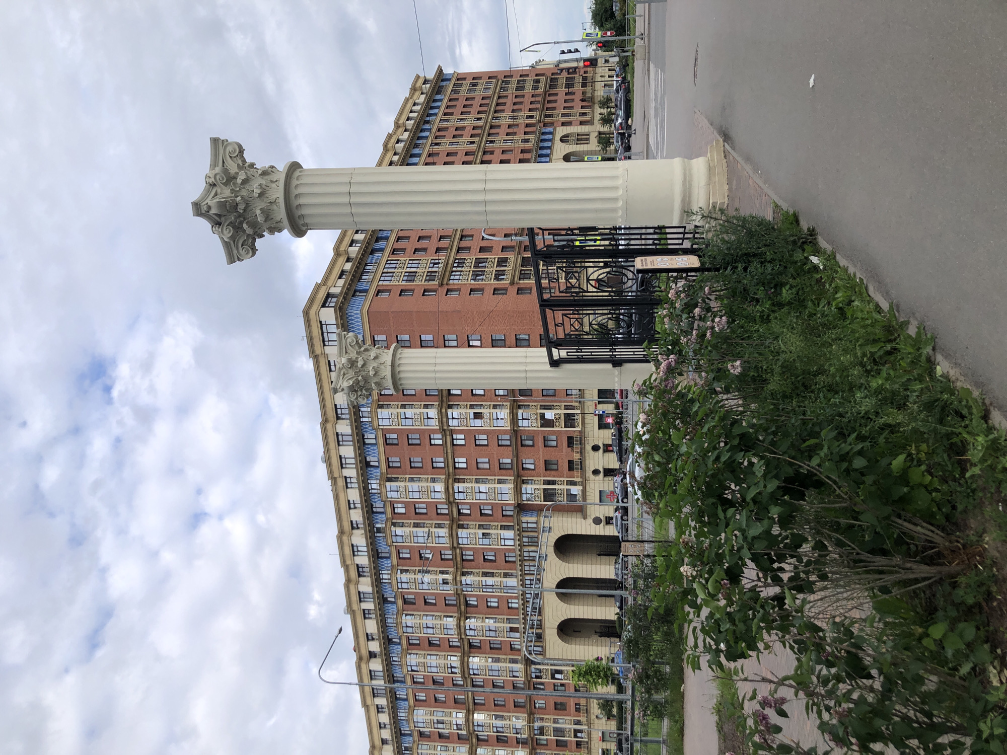 The image captures a view of a large, modern residential building with a red brick facade and numerous windows, located in Saint Petersburg, Russia. Two prominent classical columns stand in the foreground, flanking a black metal fence. Lush green bushes and flowering plants line the walkway in front of the building. Beyond the columns and fence, a street with cars and pedestrian crossings is visible. Traffic lights and streetlights are also present, indicating an urban environment. The sky is overcast with a mix of white and gray clouds. The architecture of the building suggests it is relatively new, with a blend of classical and contemporary design elements. The overall scene depicts a well-maintained urban area with residential properties and public infrastructure.