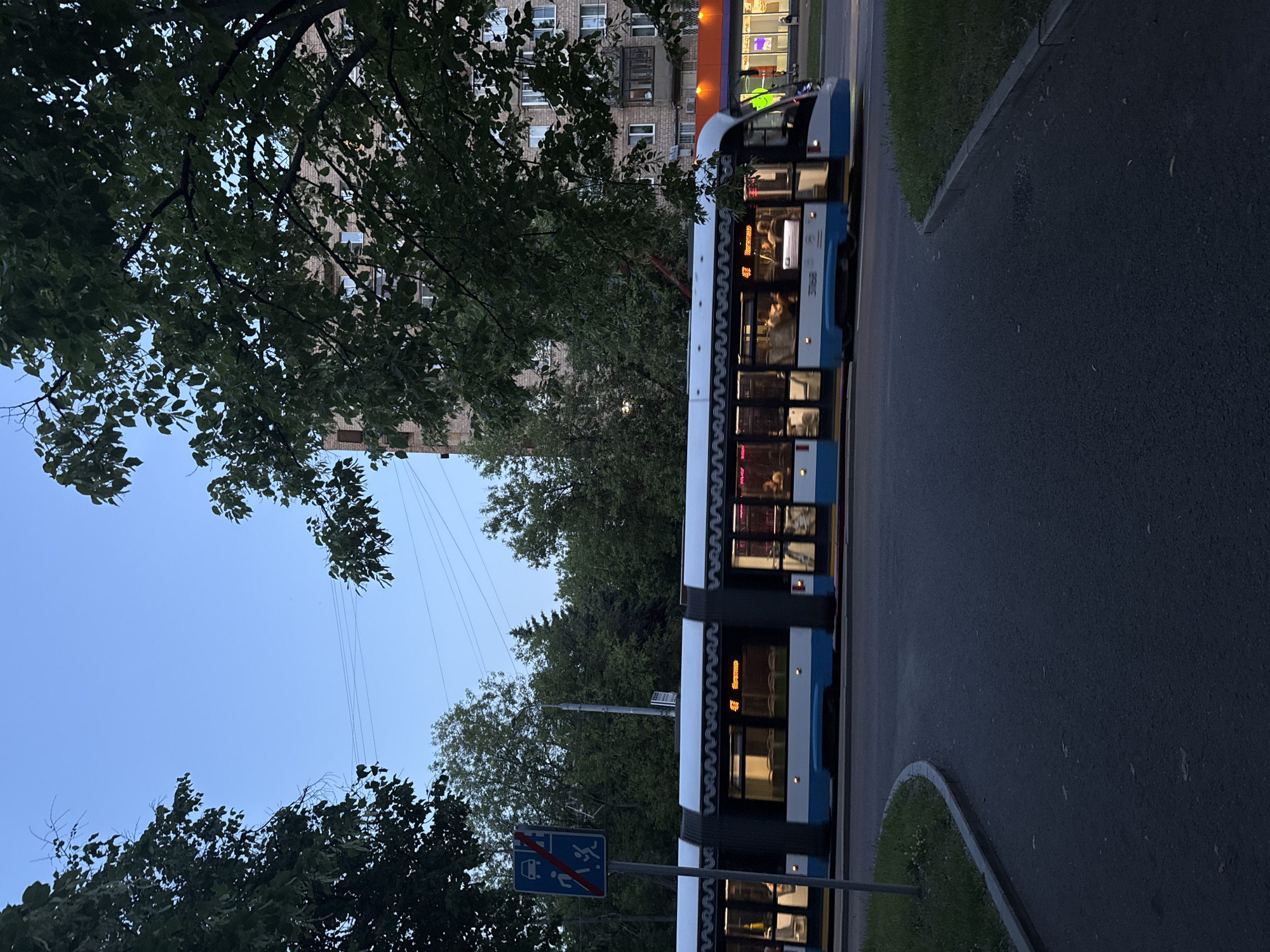 The image depicts a modern, white and blue tram moving along a street in Moscow, Russia, during the evening. The tram is a long, multi-car vehicle with illuminated windows revealing passengers inside. Its sides feature a distinctive wavy pattern along the top edge. The tram is captured in motion, indicated by a slight blur.
In the foreground, a paved road curves to the left, with a patch of grass and a curb on the left side of the frame. To the left of the tram, a blue and white traffic sign with symbols of a car and a pedestrian with a red cross over them is visible, suggesting a restriction on vehicle access and pedestrian activity in a certain area.
Behind the tram, trees with lush green foliage line the street, partially obscuring a brick apartment building. The building has multiple windows, some of which are lit, and an orange awning is visible on the ground floor of a commercial establishment further down the street. The sky above is a clear, pale blue, indicating twilight. Overhead wires are present, suggesting the tram's power source. The overall impression is a calm urban evening scene.