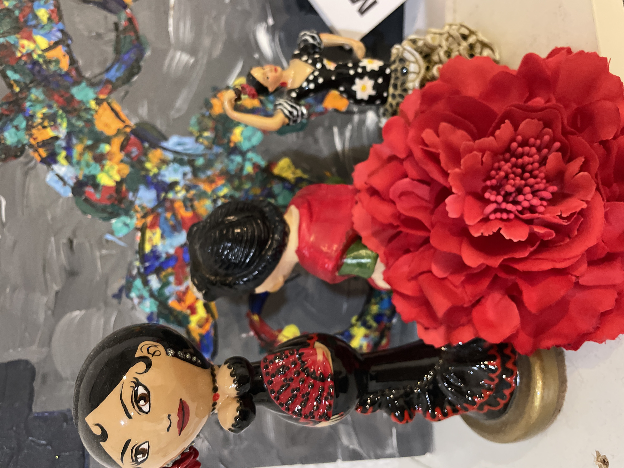 This media file presents a vibrant still life arrangement of decorative objects, likely souvenirs, displayed against an abstract painted background.
In the foreground, a prominent figurine depicts a stylized female figure, reminiscent of a flamenco dancer, with fair skin, dark eyes, red lips, and black hair. She wears an ornate black dress with red and black ruffled details and visible embellishments, possibly rhinestones, on her head. To her right, a large, artificial red flower with numerous petals and a textured pink/red stamen center dominates the right side of the image.
Further into the scene, a smaller female figurine is visible lying down. She wears a black and white polka-dot dress with white floral patterns and striped sleeves, her dark hair styled in a bun. Partially obscured behind this figure is another decorative item, appearing to be the head of an animal, possibly a leopard or cheetah, identifiable by its spotted pattern and golden hues.
All these items are positioned on a flat surface, with the background consisting of an abstract painting. This painting features a dynamic array of colorful splatters and textures in blues, greens, yellows, reds, and oranges, laid over a grey or dark grey base with lighter brushstrokes. A white rectangular object bearing partial dark text, possibly the word "NEW", is visible behind the smaller figurine.
The overall scene is a decorative display, emphasizing the aesthetic appeal of the figurines and flower. The flamenco dancer's style strongly suggests a Spanish cultural theme. No specific activity or event is taking place beyond the arrangement of these objects. The location context provided is Sochi, Russia. There is no indication of the time of day or weather.