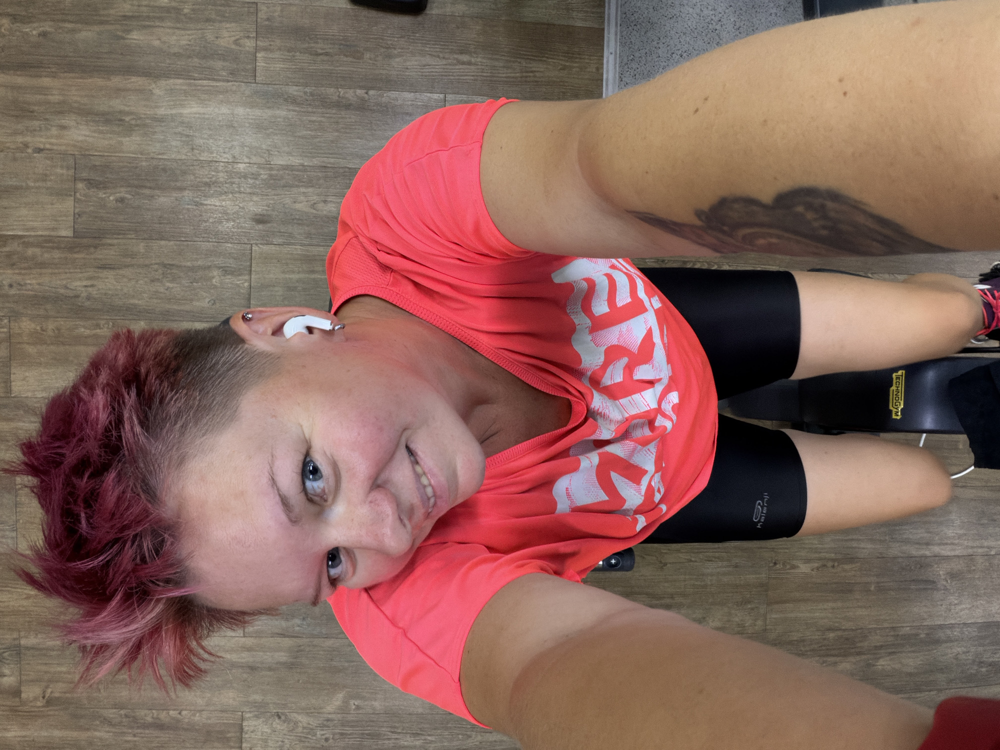 A woman with short, spiked, pink-dyed hair and blue eyes is taking a selfie. She is wearing a bright pink t-shirt with white lettering, black athletic shorts, and white wireless earbuds. She has a tattoo on her right arm. She is indoors, in what appears to be a gym, with a wooden floor and a piece of gym equipment visible. The image is taken from a high angle, looking down at the woman. There is no discernible information linking this image to the city of Chaikovsky, Russia.