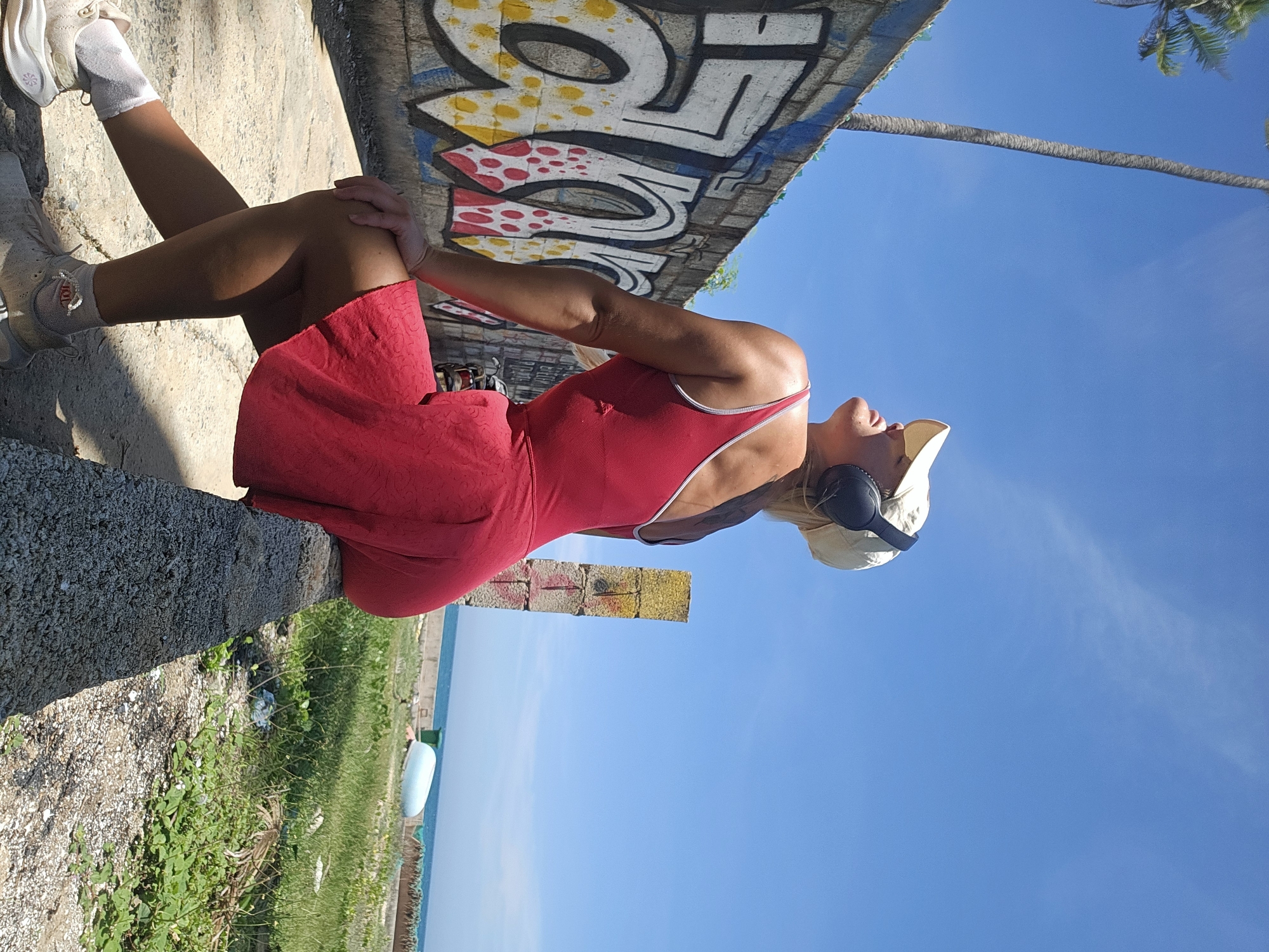 A woman with blonde hair, wearing a red outfit, a beige cap, and black headphones, is sitting on a low wall in front of a graffiti-covered wall. The wall to the left is covered in colorful graffiti. Behind the woman, a tall palm tree is visible against a clear blue sky. In the background, there is a body of water, green grass, and a small blue boat. The scene appears to be outdoors, possibly in a coastal area of Phu Thuy Ward, Vietnam, given the presence of palm trees and the ocean. The woman is looking upwards with her eyes closed, suggesting she is enjoying the moment or listening to music.