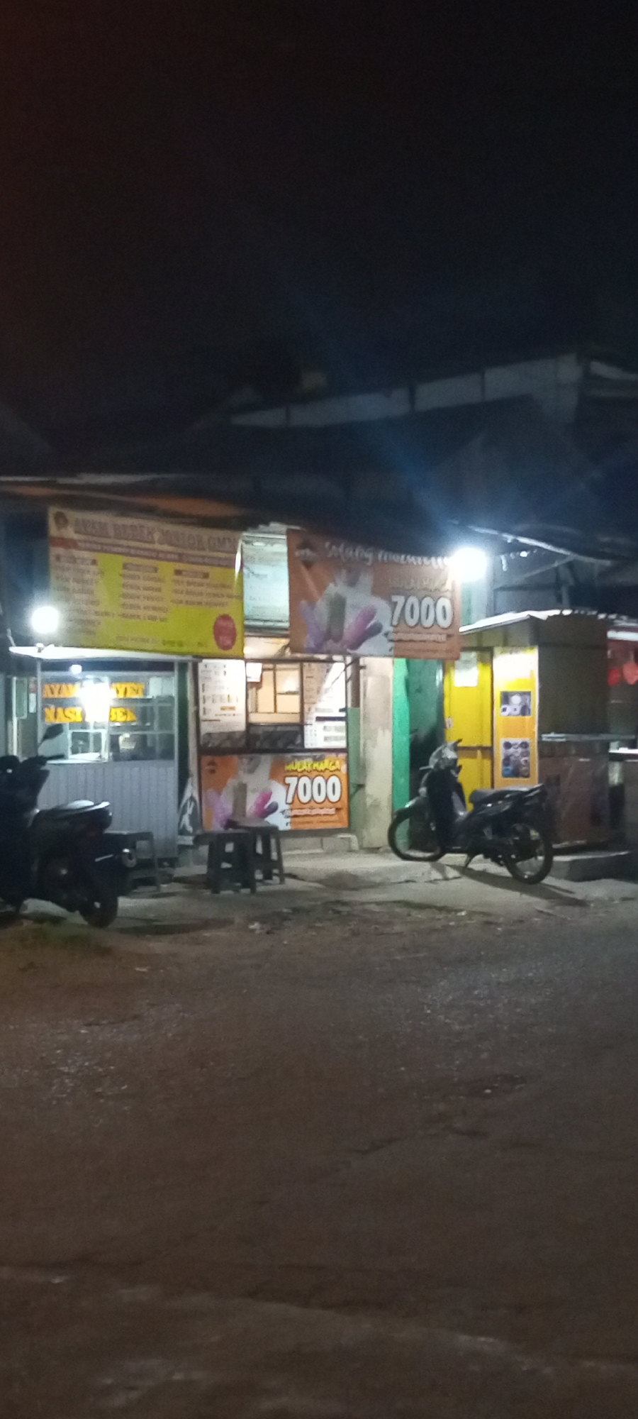 The image depicts a street scene at night in Setu, Indonesia, likely an area with food stalls or small businesses. In the foreground, the asphalt road is visible, showing some texture and reflections. To the left, a black motorcycle is parked, partially obscuring a storefront with a glass window and what appears to be food items displayed inside. Above the storefront, a yellow banner with text, possibly a menu or advertisement, is hung.
In the center of the image, several dark plastic stools are arranged in front of another stall. A prominent orange banner with text and an image of what looks like a drink or food item is displayed here, with the number "7000" clearly visible, likely indicating a price. Further to the right, another motorcycle is parked. Behind the stalls, the structures appear to be made of corrugated metal and possibly wood, suggesting a temporary or semi-permanent setup common in street markets. The lighting is dim, with artificial lights from the stalls casting a warm glow, creating shadows and highlighting certain elements of the scene. The overall impression is a busy, informal commercial area at night.