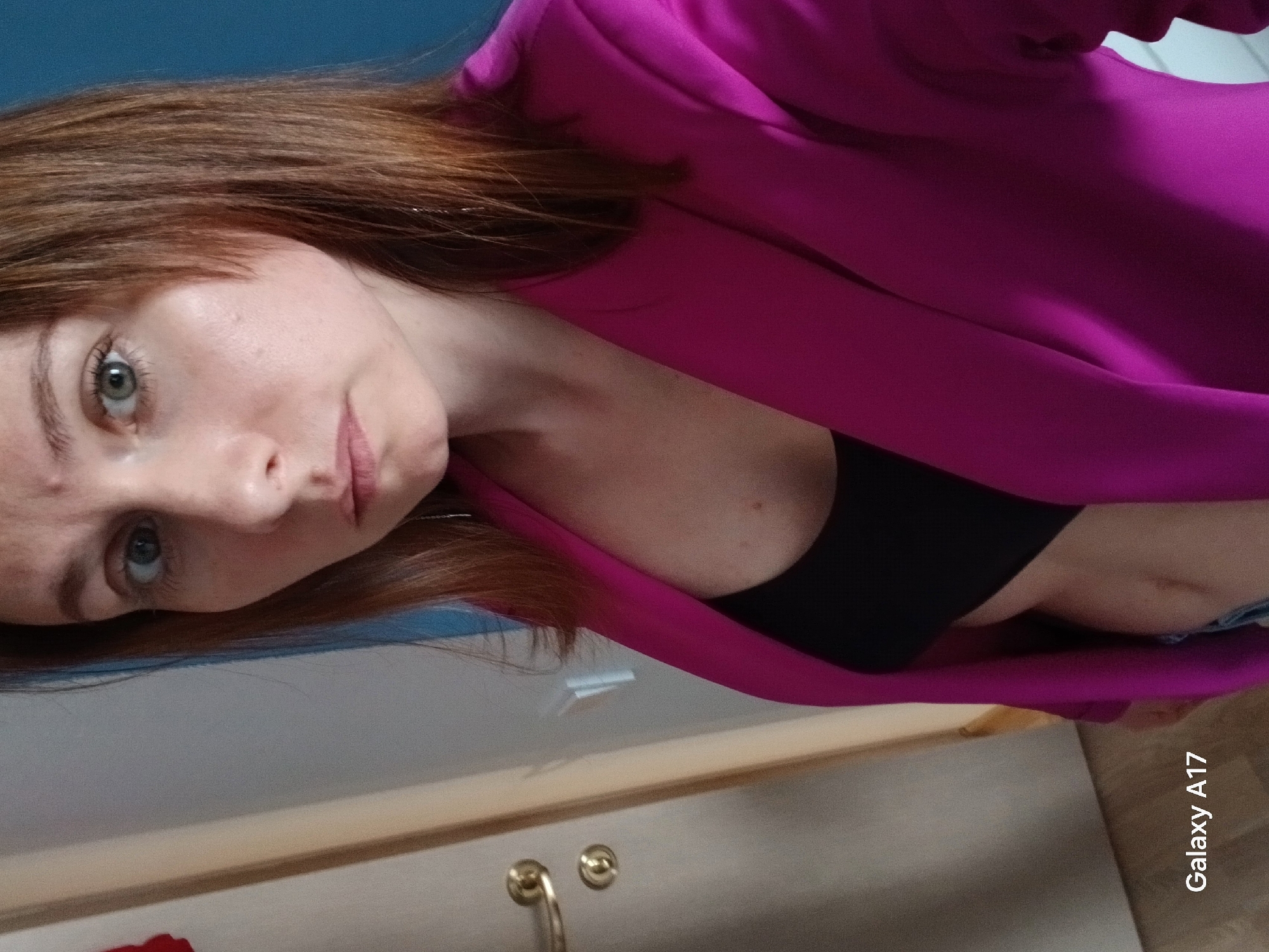 A woman with light skin, auburn hair, and blue eyes is wearing a black top and a bright pink blazer. The photo appears to be taken indoors. The background includes a blue wall, a white surface with golden drawer pulls, and a section of wooden floor. The orientation of the image is sideways. The phone model "Galaxy A17" is visible in the bottom right corner. There is no information to indicate a specific location within Yekaterinburg, Russia.