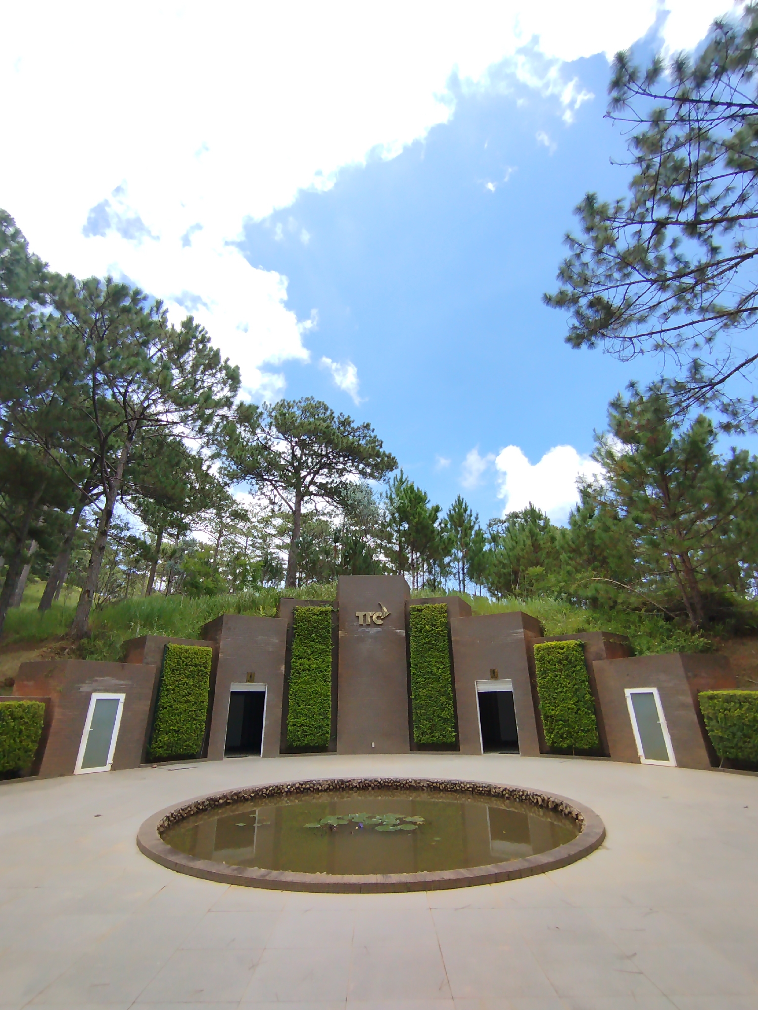 The image depicts a serene outdoor scene at the entrance of a building, likely a tourist attraction or a memorial, situated in Lam Vien - Da Lat, Vietnam. The architecture is modern and striking, featuring a central concrete structure adorned with vertical green hedges on either side of two doorways. The building's facade is a dark, possibly brown or grey, concrete with angular designs. To the left and right of the main structure, there are two identical, narrow, rectangular white doors set against the same concrete. A circular pond with lily pads occupies the foreground, bordered by a stone rim and surrounded by a light-colored tiled walkway. Pine trees and lush green vegetation are visible on the hillside behind the building, contributing to the natural beauty of the location. The sky above is a clear blue with scattered white clouds, suggesting a pleasant day. The "TTC" logo is prominently displayed on the central part of the building. There are no people or animals visible in the image. The overall atmosphere is one of tranquility and sophisticated design integrated with nature.