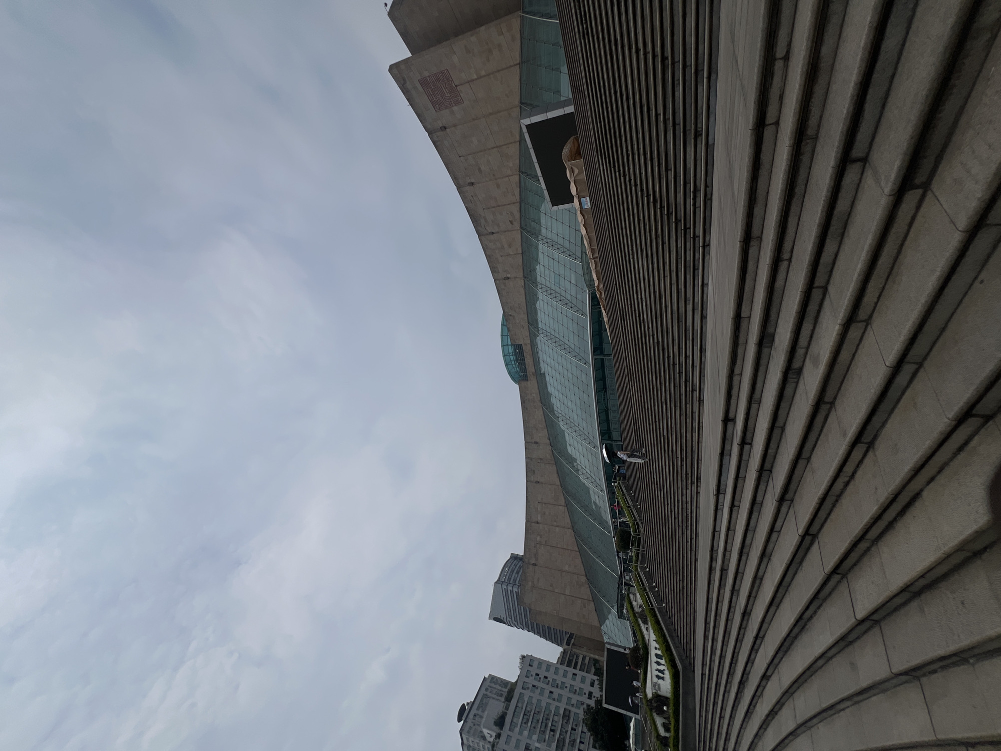 A wide-angle, low-angle outdoor photograph captures the exterior of a modern, curved building with a glass facade and a large, stepped entrance in Yuzhong District, China. The building's facade is made of large concrete slabs and extensive glass panels. The foreground is dominated by a vast, multi-tiered staircase, with curved sections at the bottom and straight sections leading up to the building's entrance. Several individuals are visible on the stairs. One person, dressed in light-colored clothing, stands with an umbrella on the middle section of the stairs, facing away from the camera. Other people are scattered further up the stairs, some walking and some standing, also appearing to be visitors. On the left side of the frame, a row of potted plants and a low wall with sculpted figures are visible. In the background, several tall, modern buildings of various architectural styles rise into a cloudy sky. The overall scene depicts a public space with grand architectural features and a moderate number of people engaged in accessing the building.