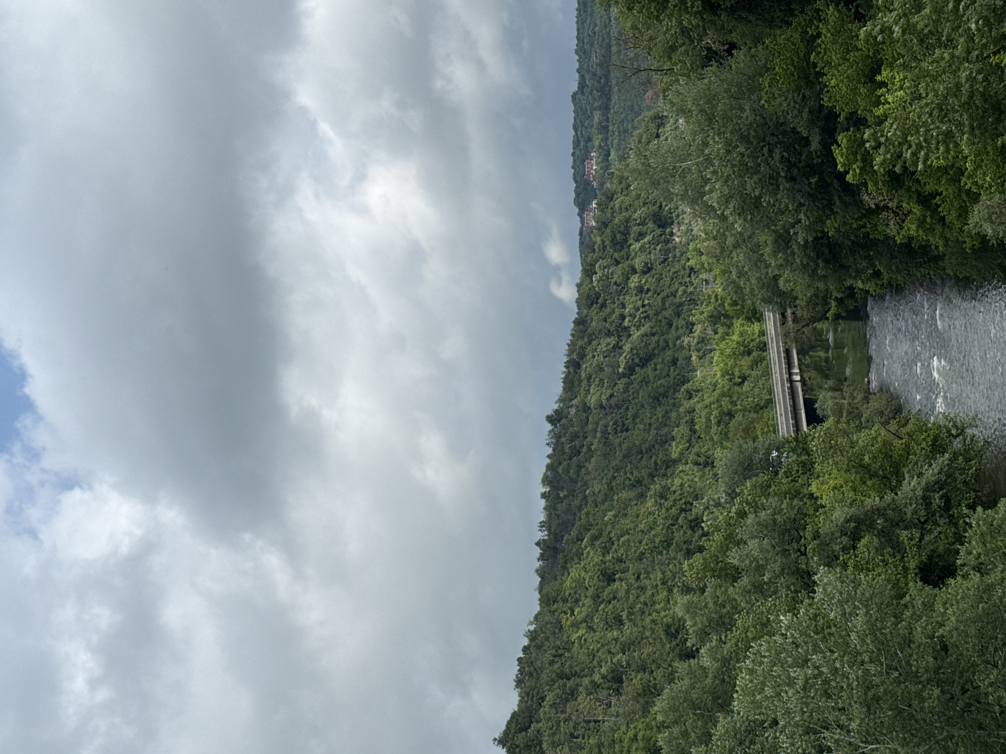 The image depicts a landscape in Veliko Tarnovo, Bulgaria, featuring a river flowing between forested hills. The sky is overcast with heavy clouds. A bridge spans the river, partially obscured by trees. A small village can be seen on the distant hillside. The trees are lush and green, indicating a natural setting.